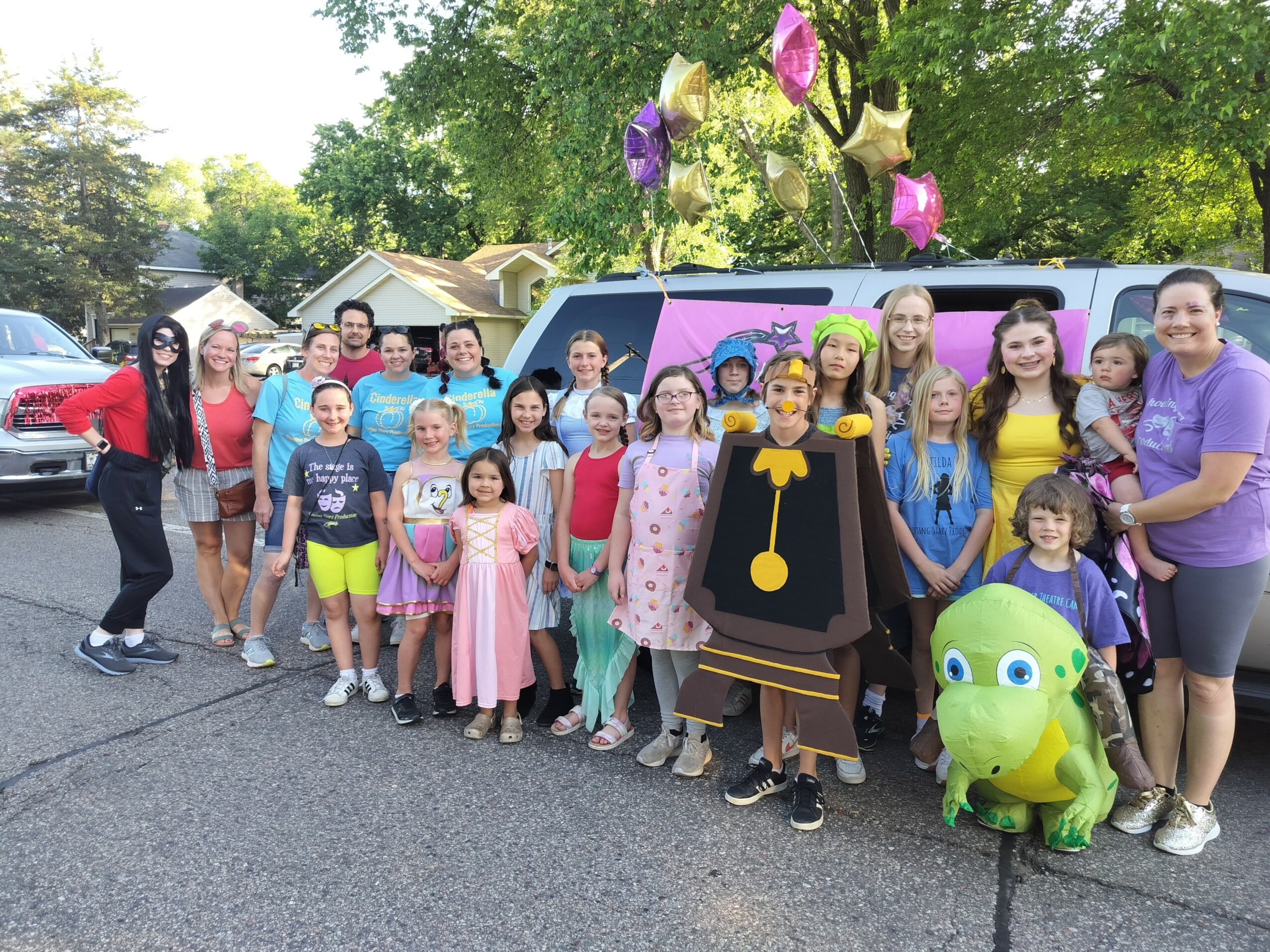 Group of children and adults outdoors, some in costumes, with a decorated vehicle and balloons in the background.