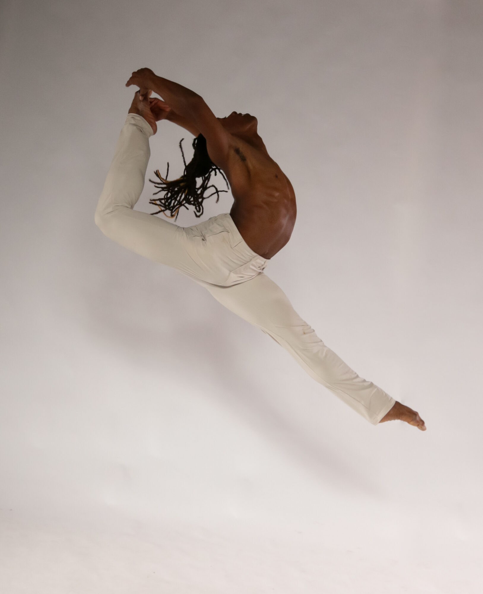 Person with dreadlocks performs a leap with legs extended and arms reaching back, against a plain background.