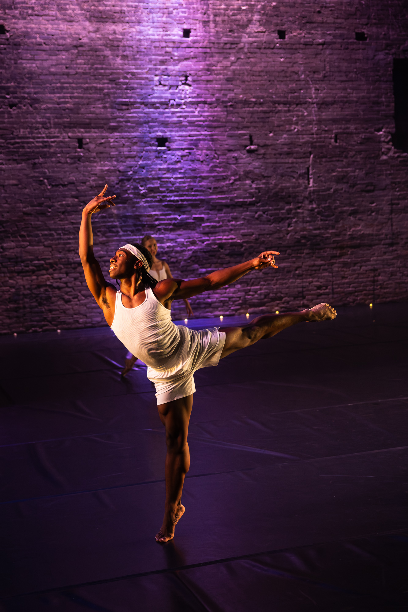 A dancer performs a pose on stage with a brick wall background illuminated in purple light.