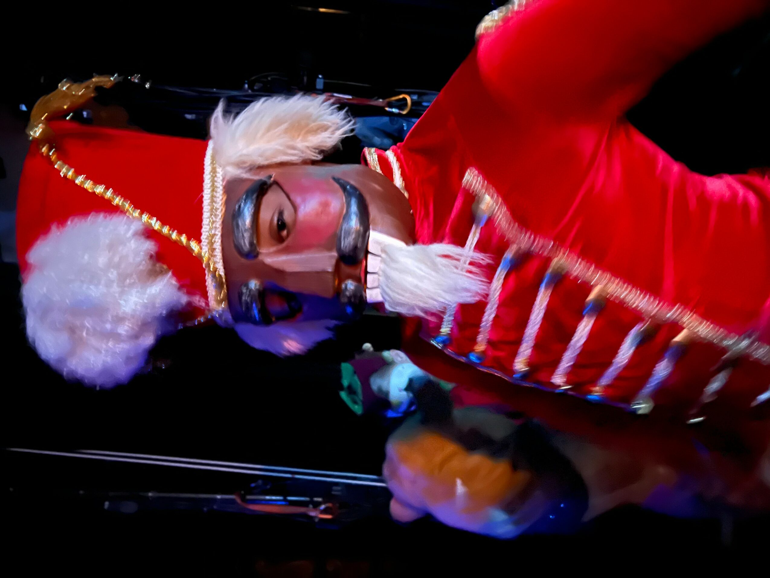 Person dressed as a Christmas elf with a Santa hat, glasses, and a red outfit, holding a small object, against a dark background.