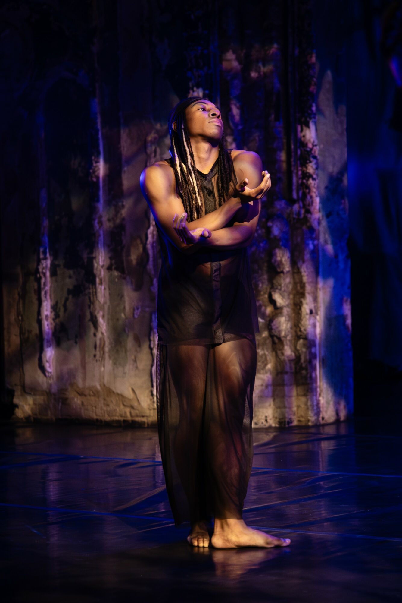 A woman stands on stage with arms crossed, wearing a sheer black dress, in front of a textured backdrop, under stage lighting.