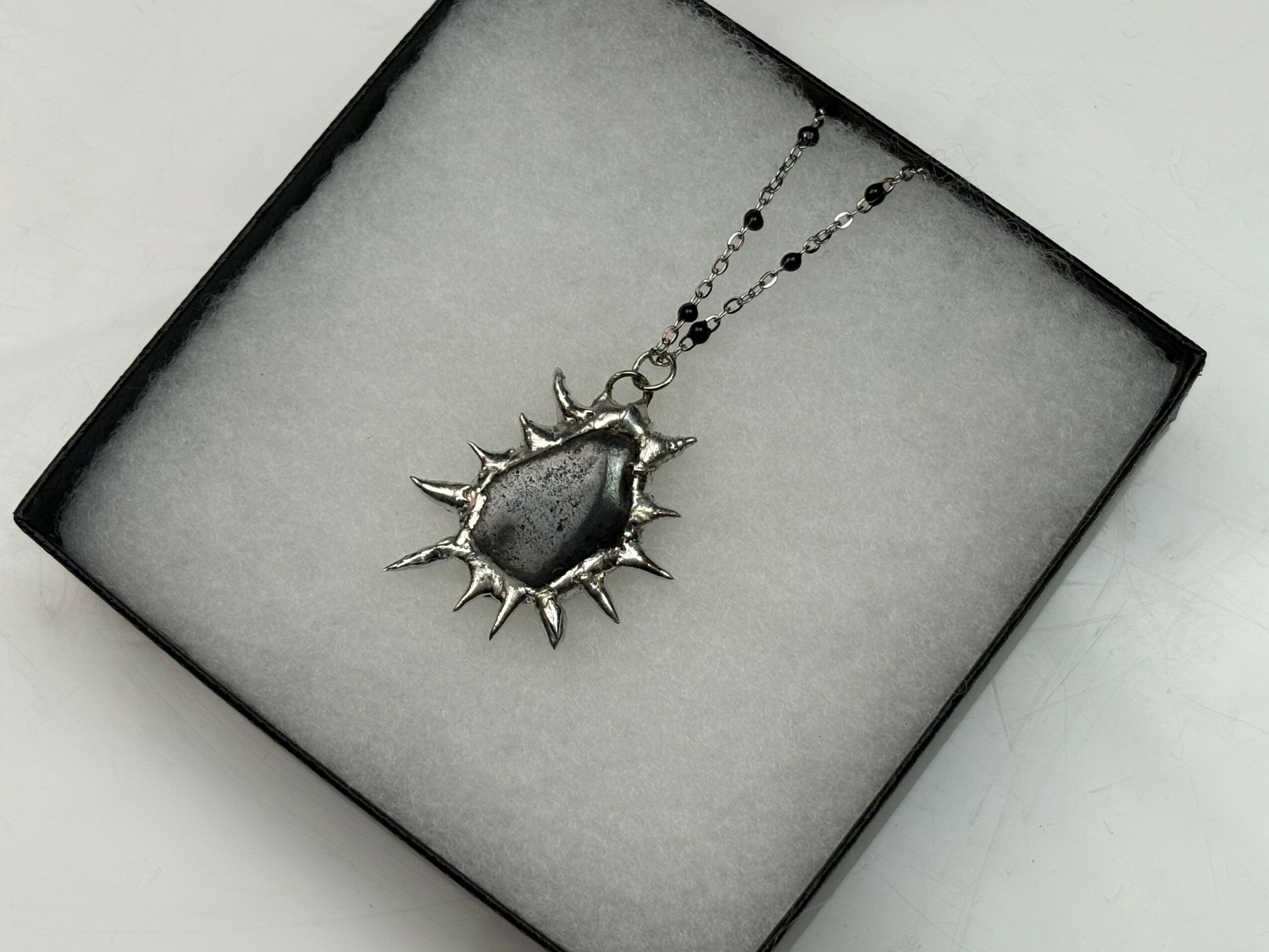 Silver necklace with a large, spiked, sun-shaped pendant inside a black box with white padding.