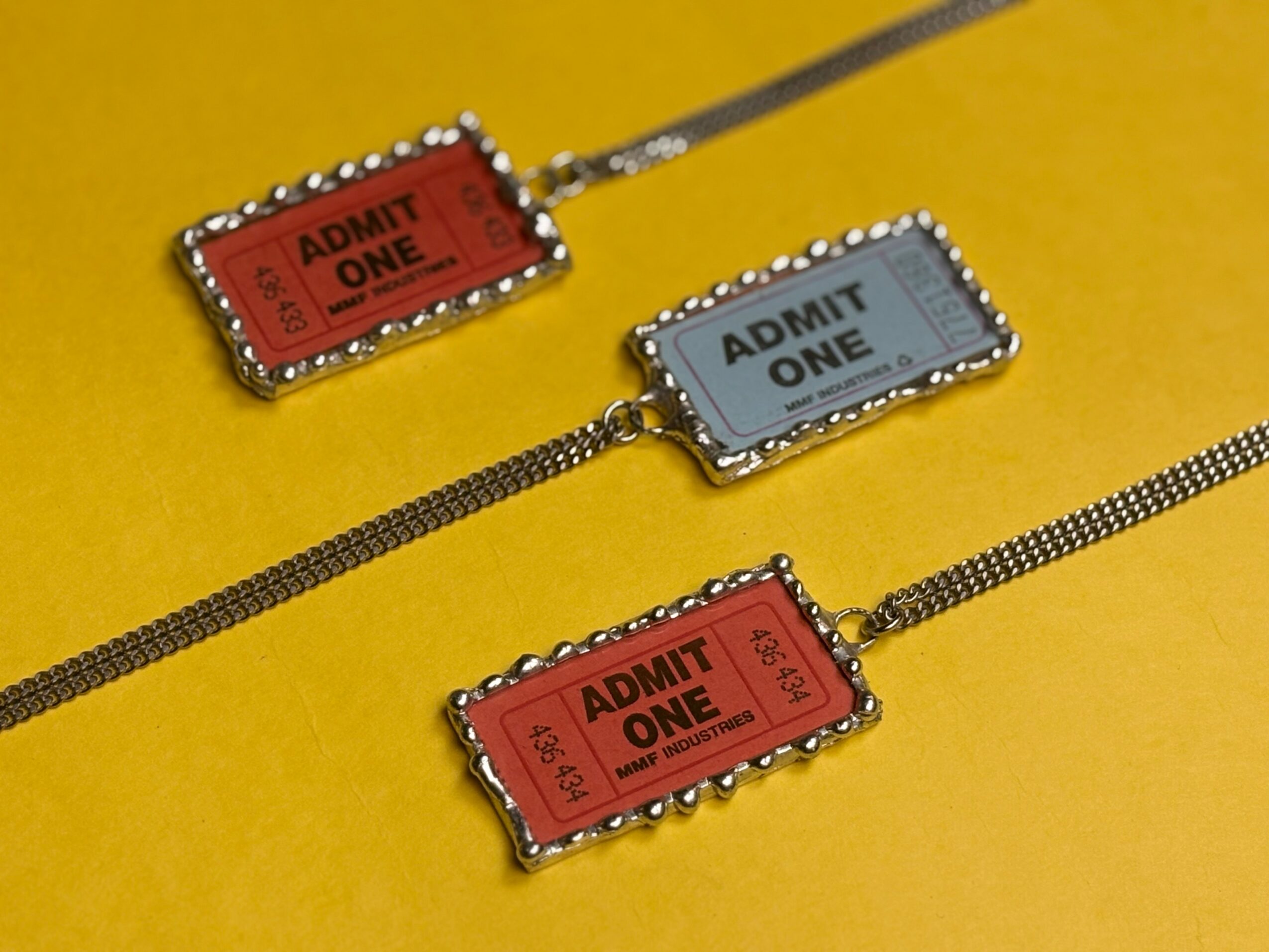 Three jewelry pieces with ticket-shaped charms, two red and one gray, on a yellow background.