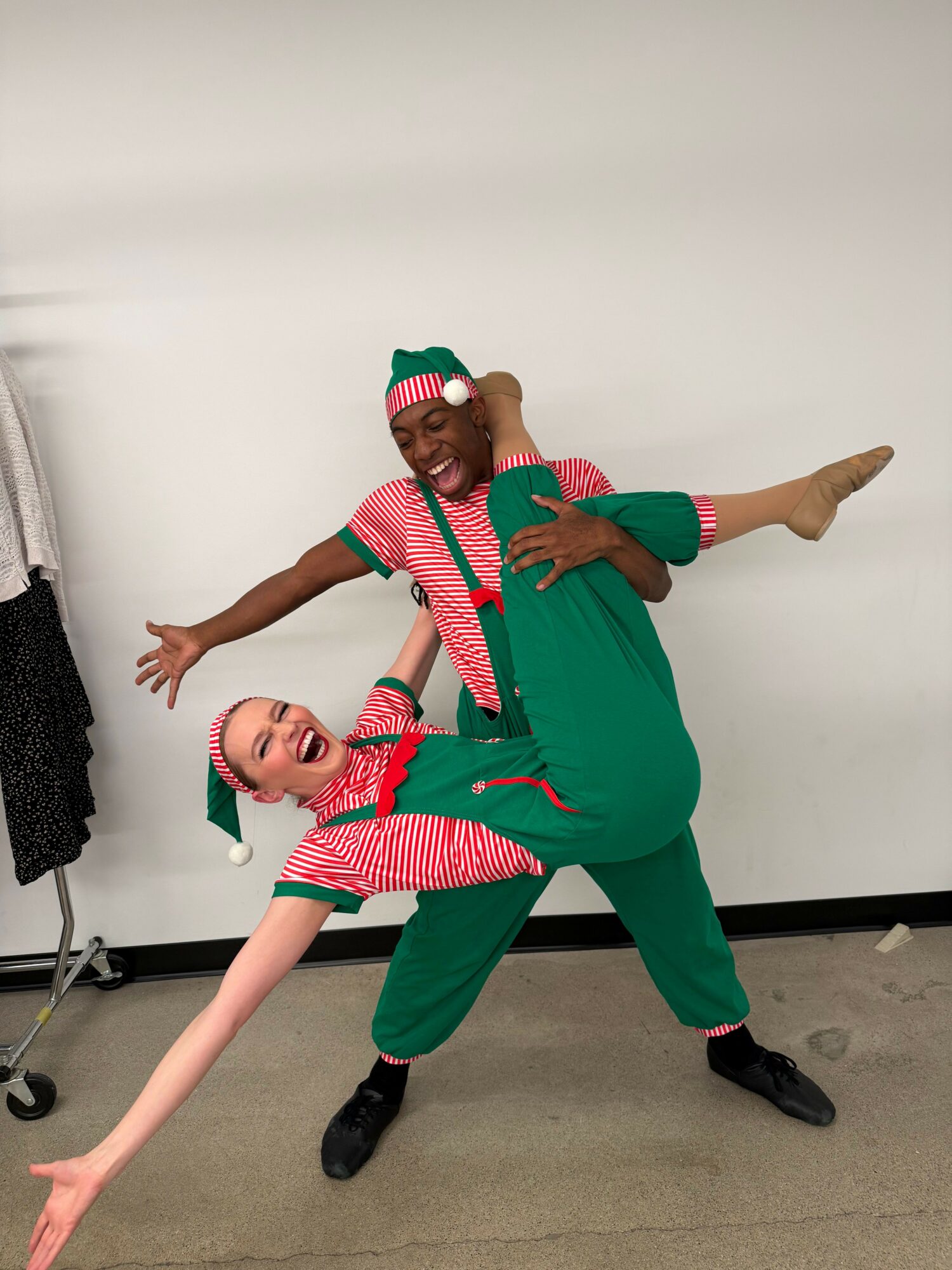 Two people dressed in festive Christmas costumes pose playfully against a plain white background, one lying on the floor and the other leaning over.