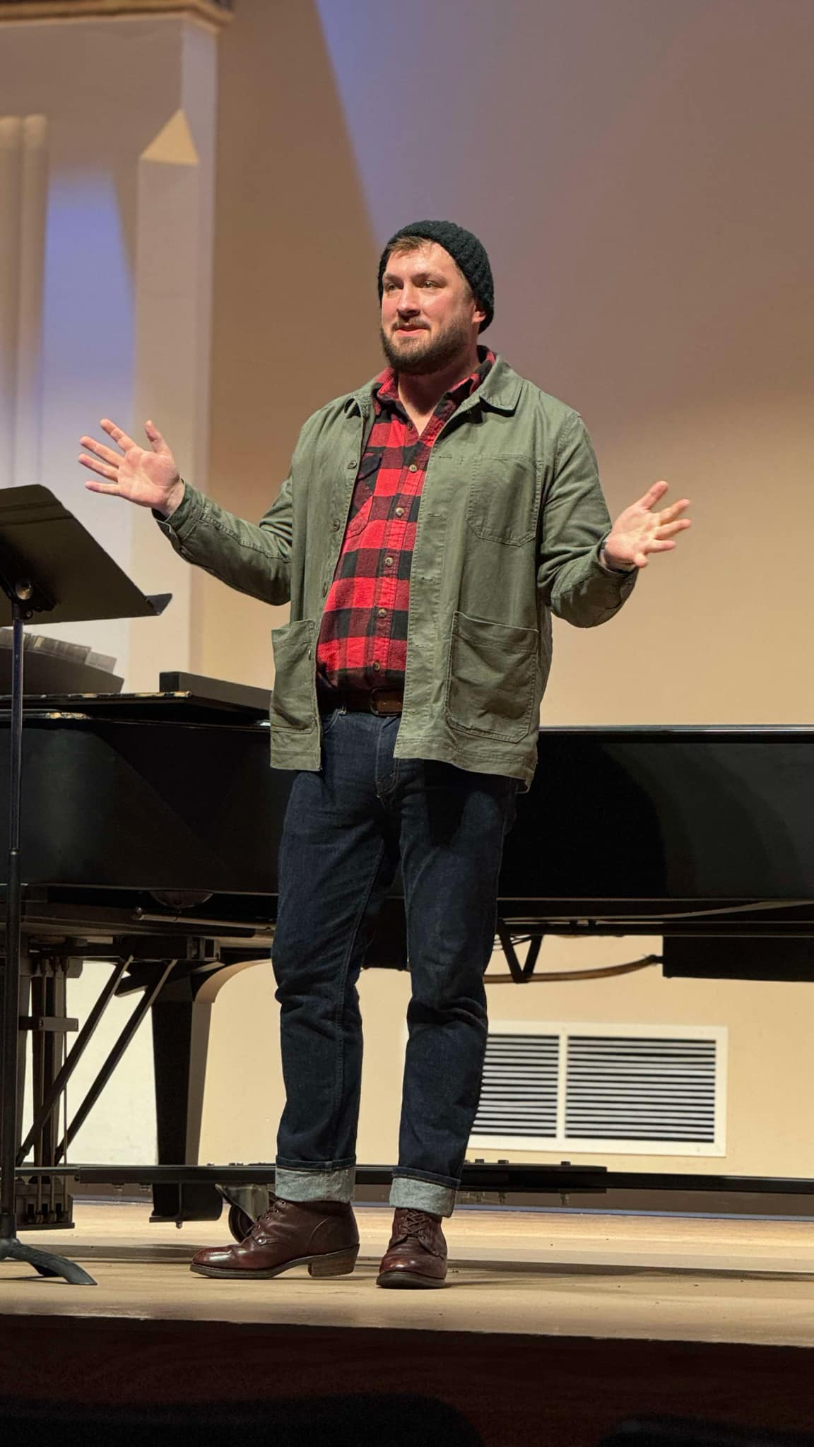 Person standing on stage with arms outstretched, wearing a green jacket, red plaid shirt, and dark pants, in front of a piano.