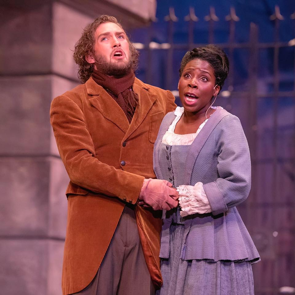 Two actors on stage, one man with curly hair and beard, wearing a brown coat, and one woman with short hair, wearing a gray dress, holding hands and looking upward.
