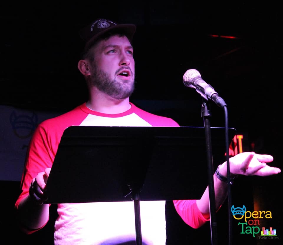 Man with beard and cap speaking into a microphone on stage, holding a hand out, with a music stand in front.