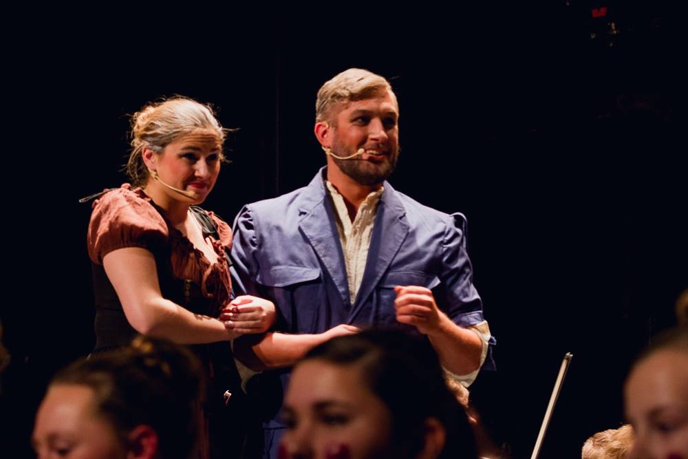 Two people on stage, a woman and a man, standing close together, with a dark background. The woman has blonde hair and wears a brown dress. The man has short hair, a beard, and wears a blue jacket. They are holding hands and looking at the audience.