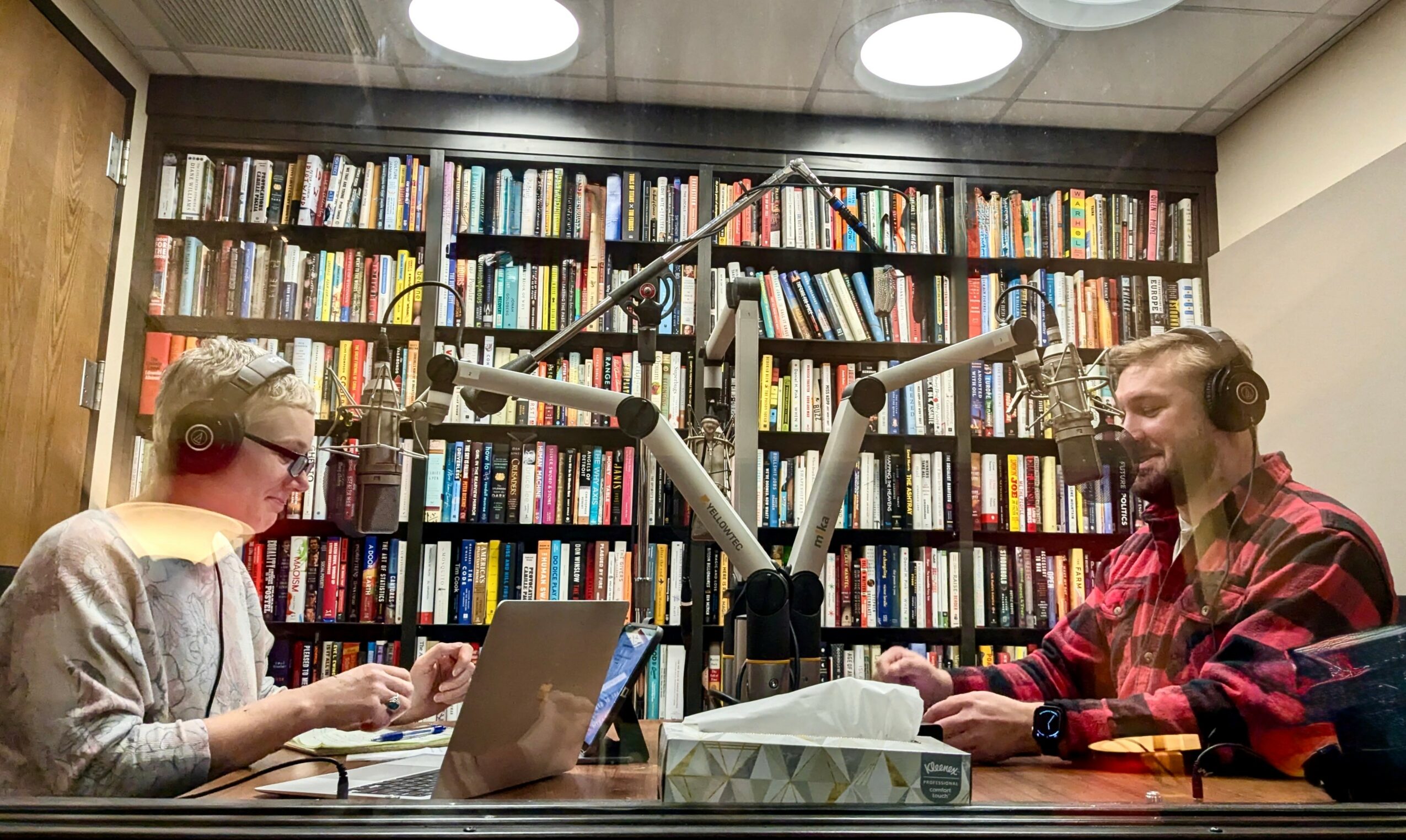 Two people sit at a table in a library with a large bookshelf filled with books behind them, speaking into microphones.