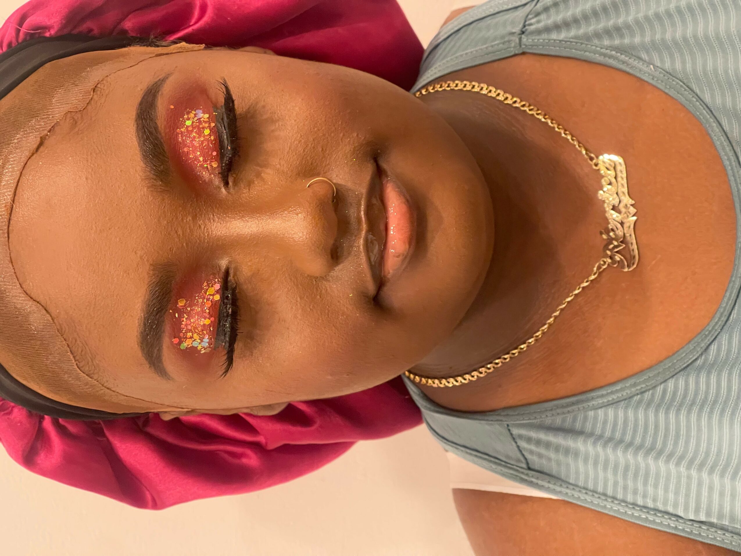 Close-up of a woman with closed eyes, makeup, gold necklace, and a pink headscarf, lying down.