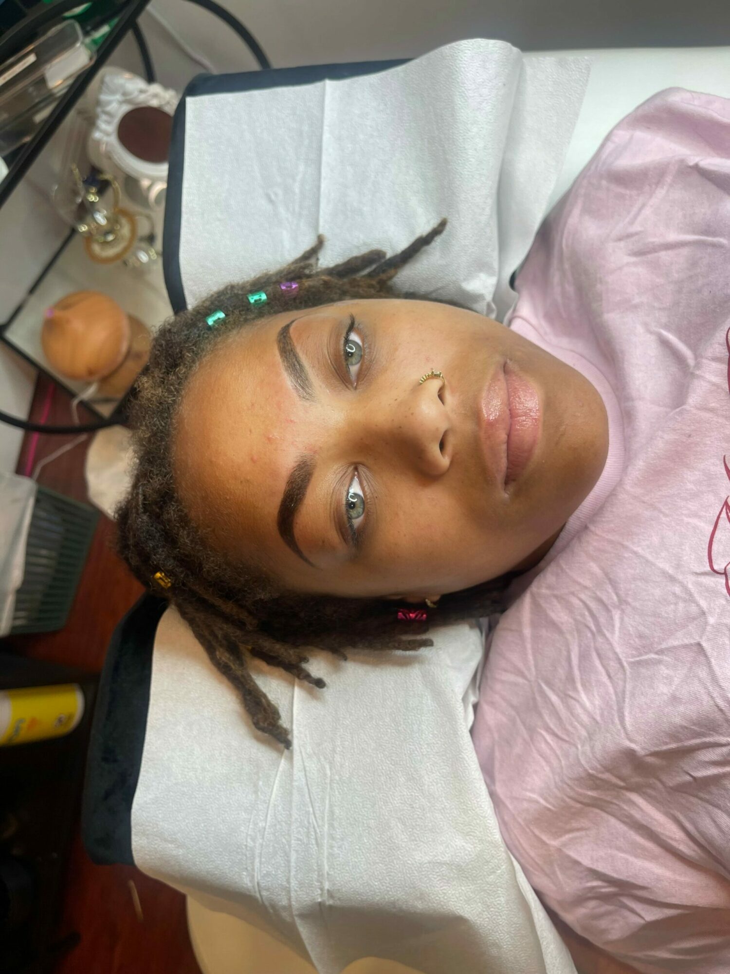 Person lying on a treatment table with eyes closed, head tilted, and colorful beads in hair, in a medical setting.
