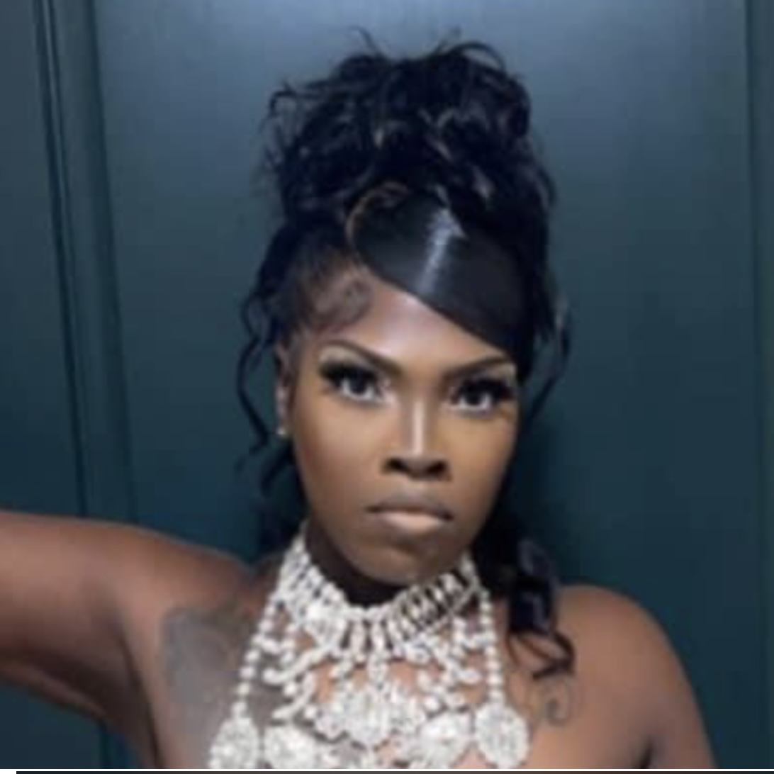 Woman with styled hair, wearing multiple pearl necklaces, looking directly at the camera, dark background.