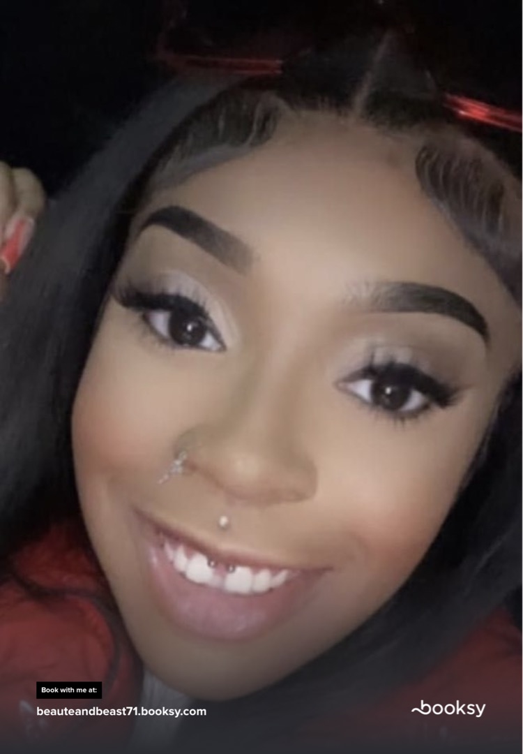 Close-up of a smiling young woman with dark hair, makeup, and facial piercings, in a dark background.