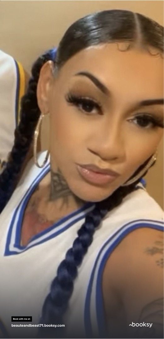 Close-up of a woman with braided hair, makeup, and tattoos, wearing a sports jersey, looking at the camera.