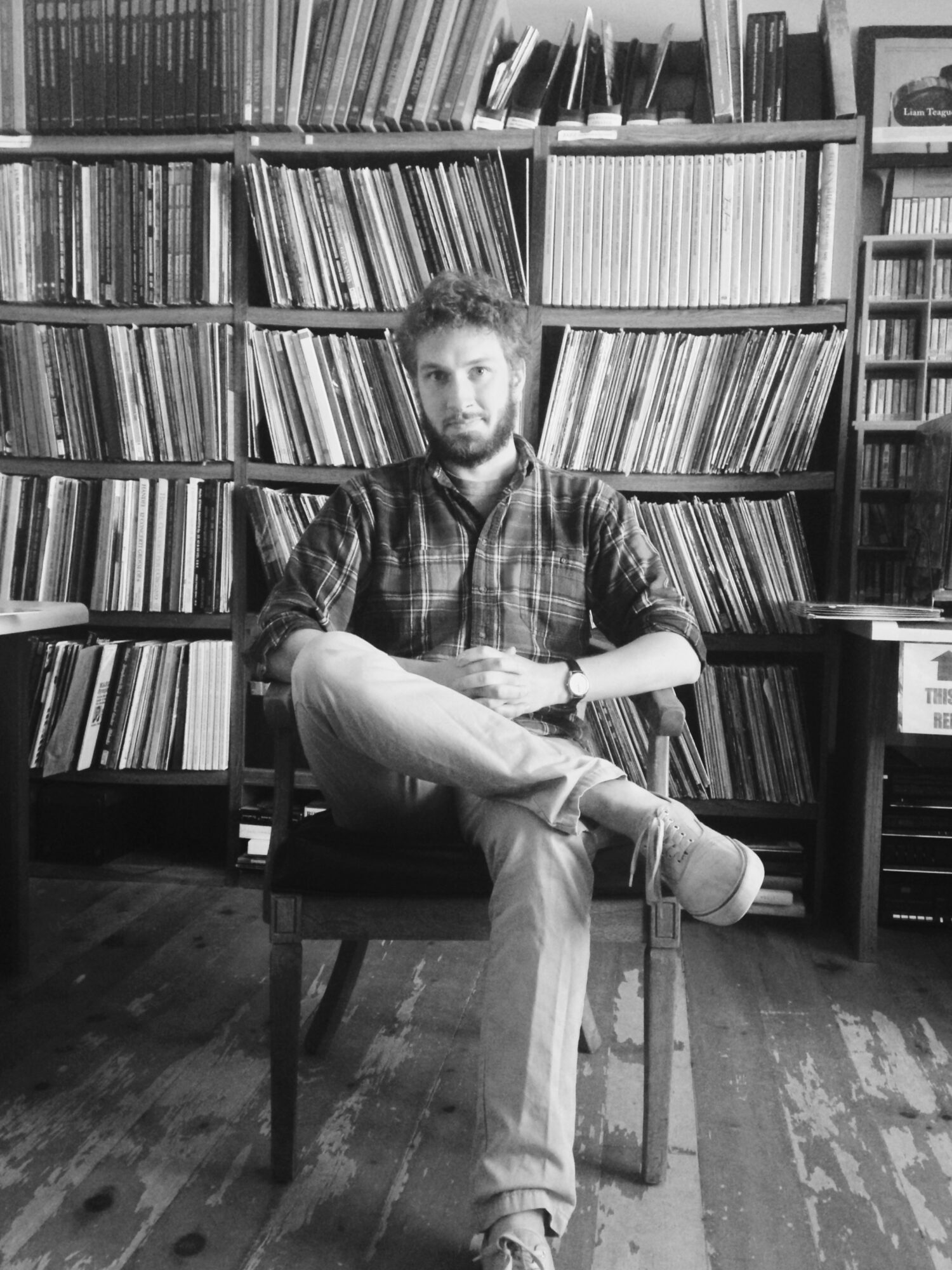 Man with beard sitting on a chair in front of bookshelves filled with books, in a room with wooden floor.