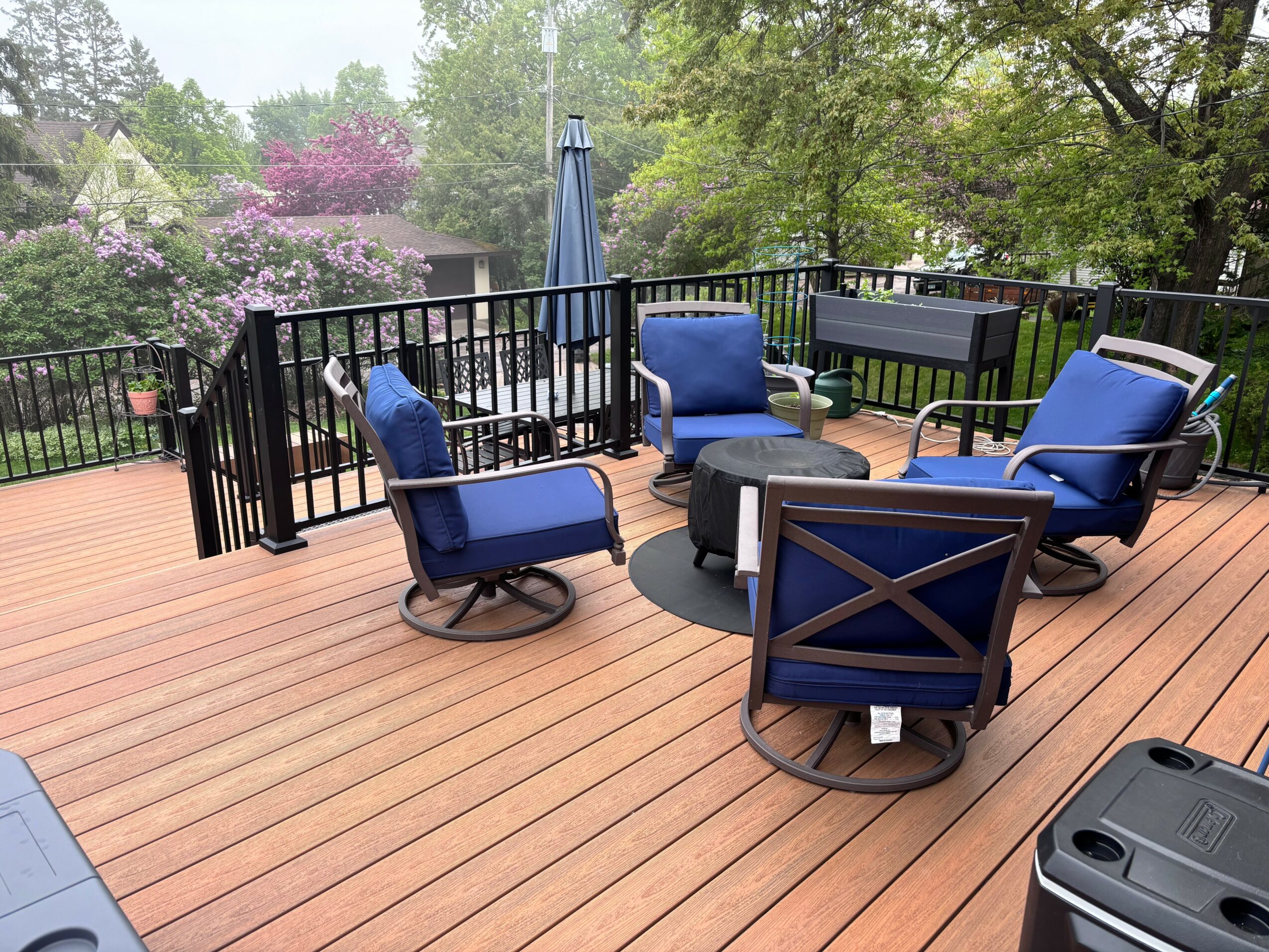 Outdoor deck with blue cushioned chairs, a round table, and a closed umbrella, surrounded by trees and flowering plants.