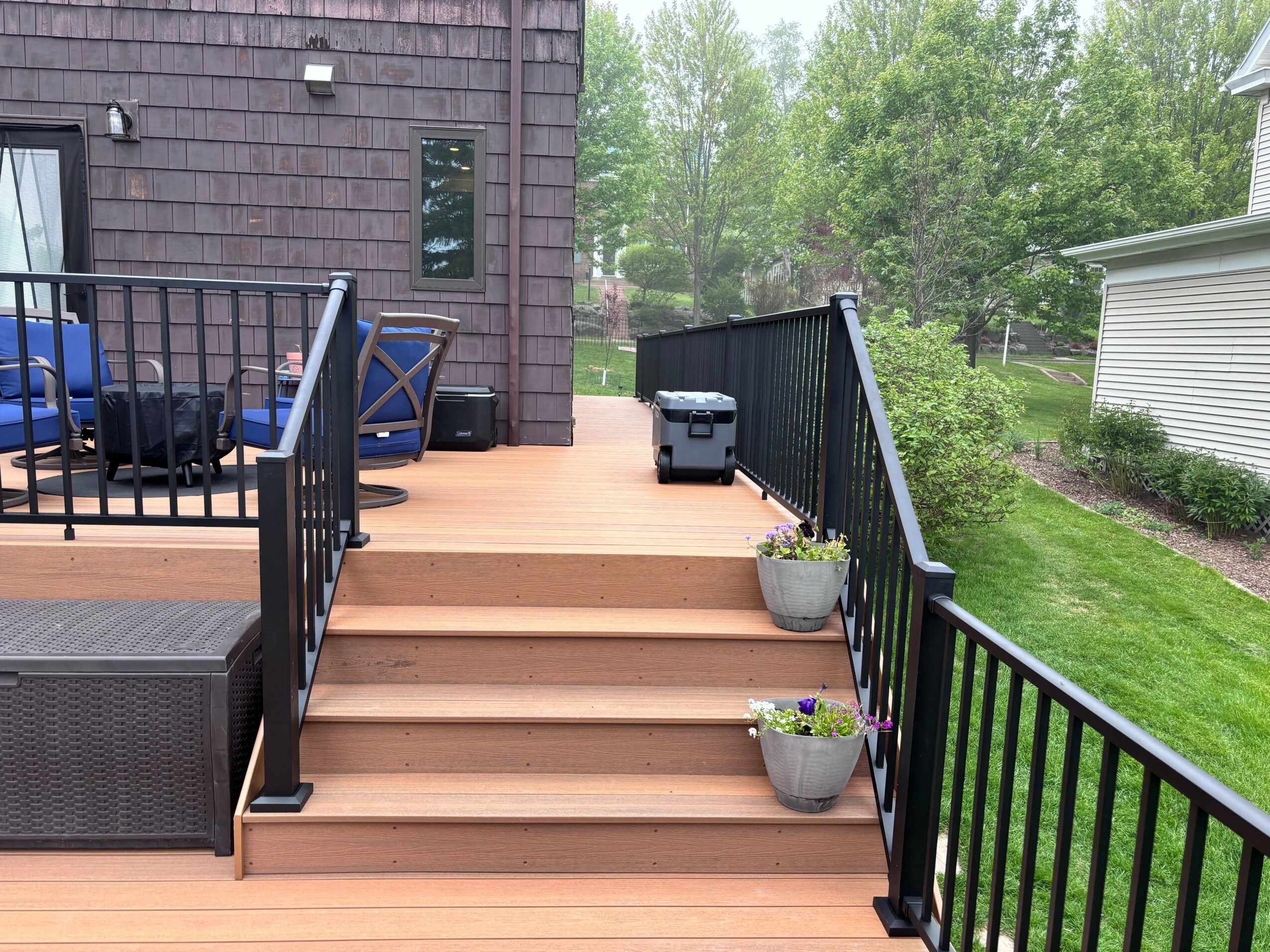 Wooden deck with stairs, black railing, potted plants, and outdoor furniture, surrounded by green trees and grass.