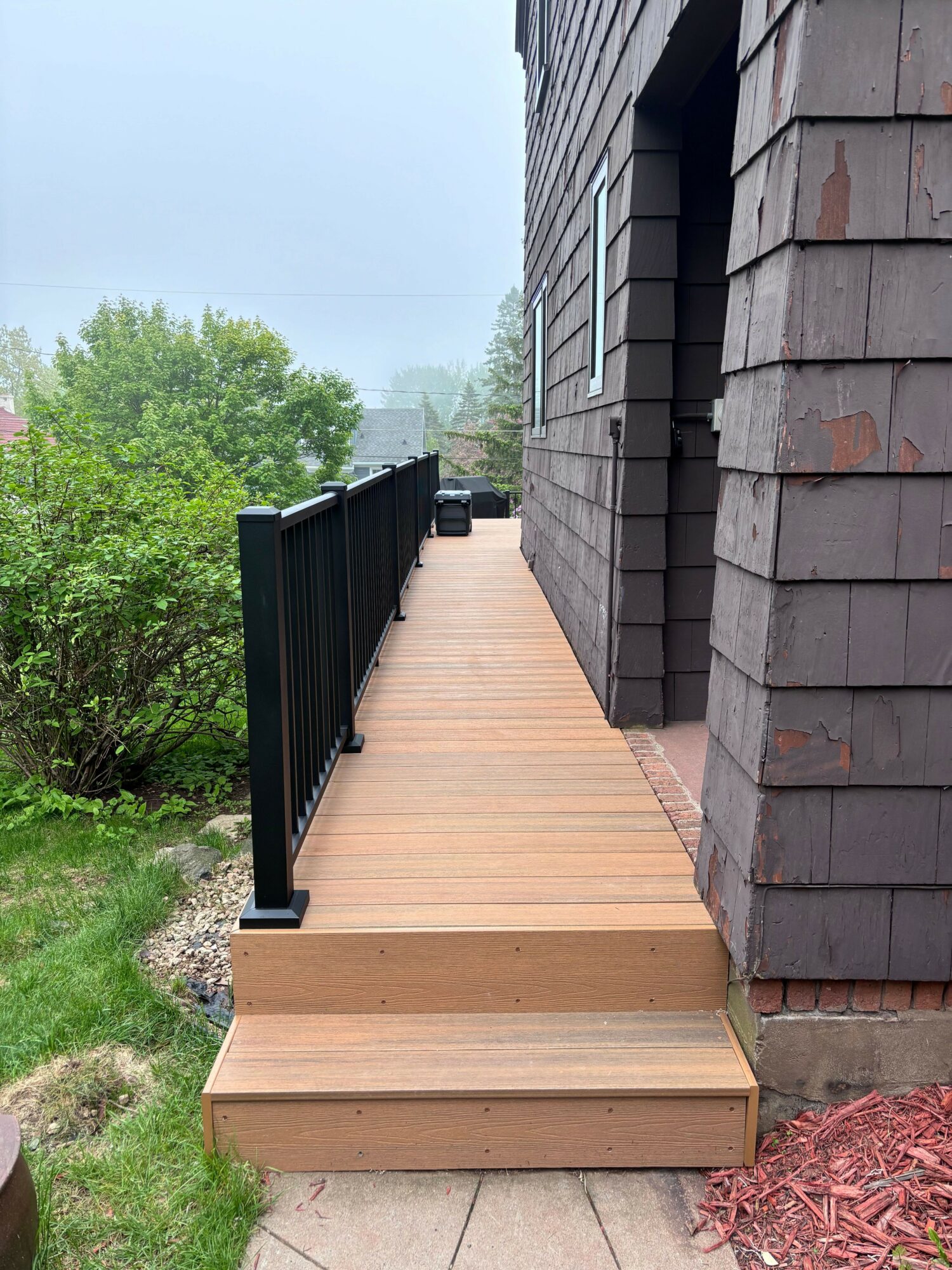 Wooden ramp with black railing leading to a brick building, surrounded by greenery and pink mulch.