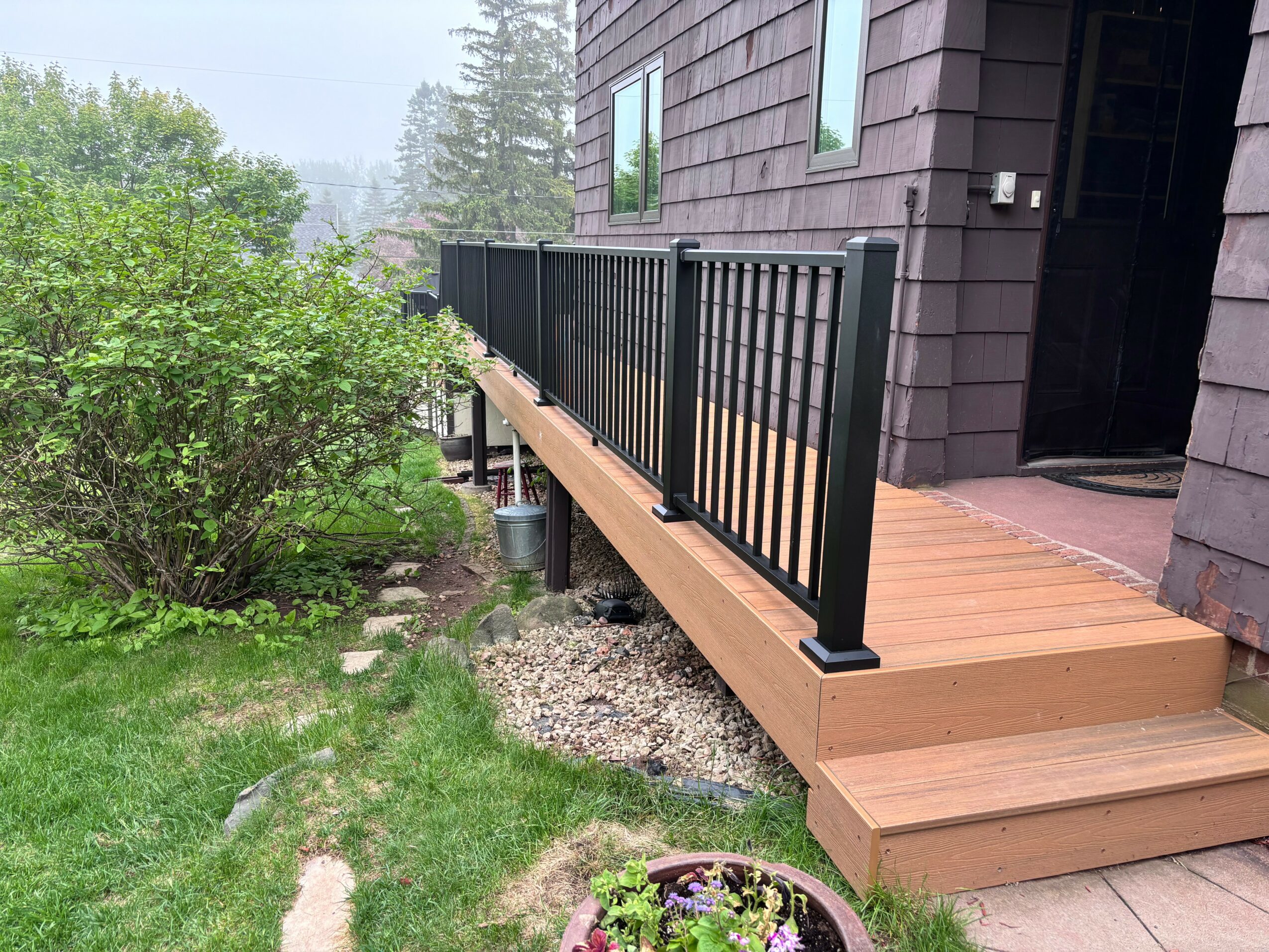 Wooden deck with black railing attached to a dark-colored house, surrounded by green grass and bushes.