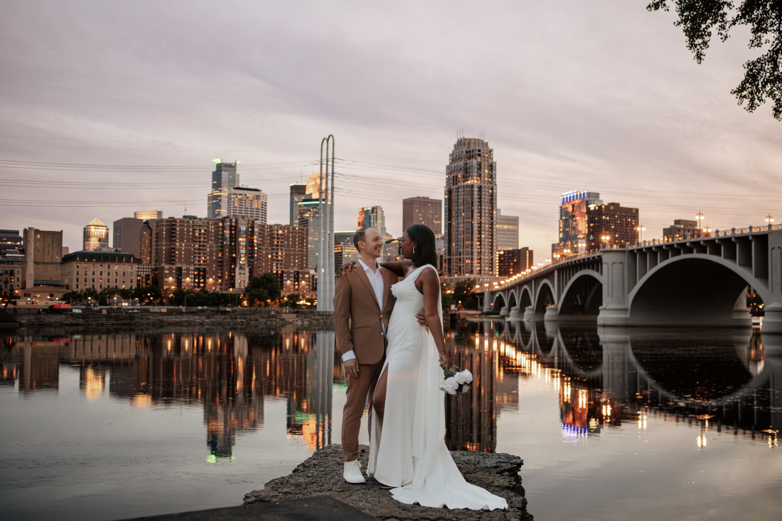 A couple in wedding attire stands by a river with a city skyline and bridge in the background.