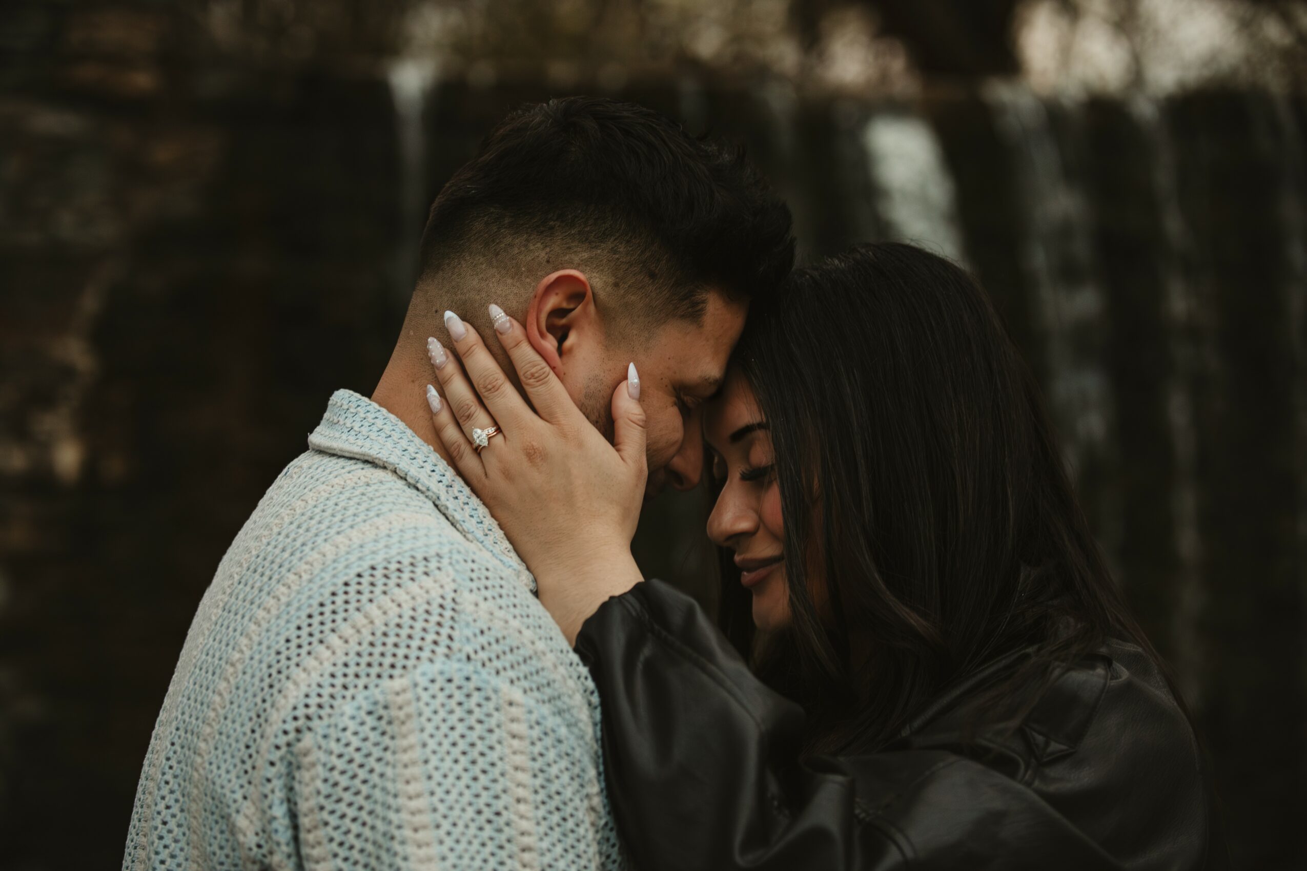 Two people with their foreheads touching, one with hand on the other's face, in an outdoor setting with a waterfall background.
