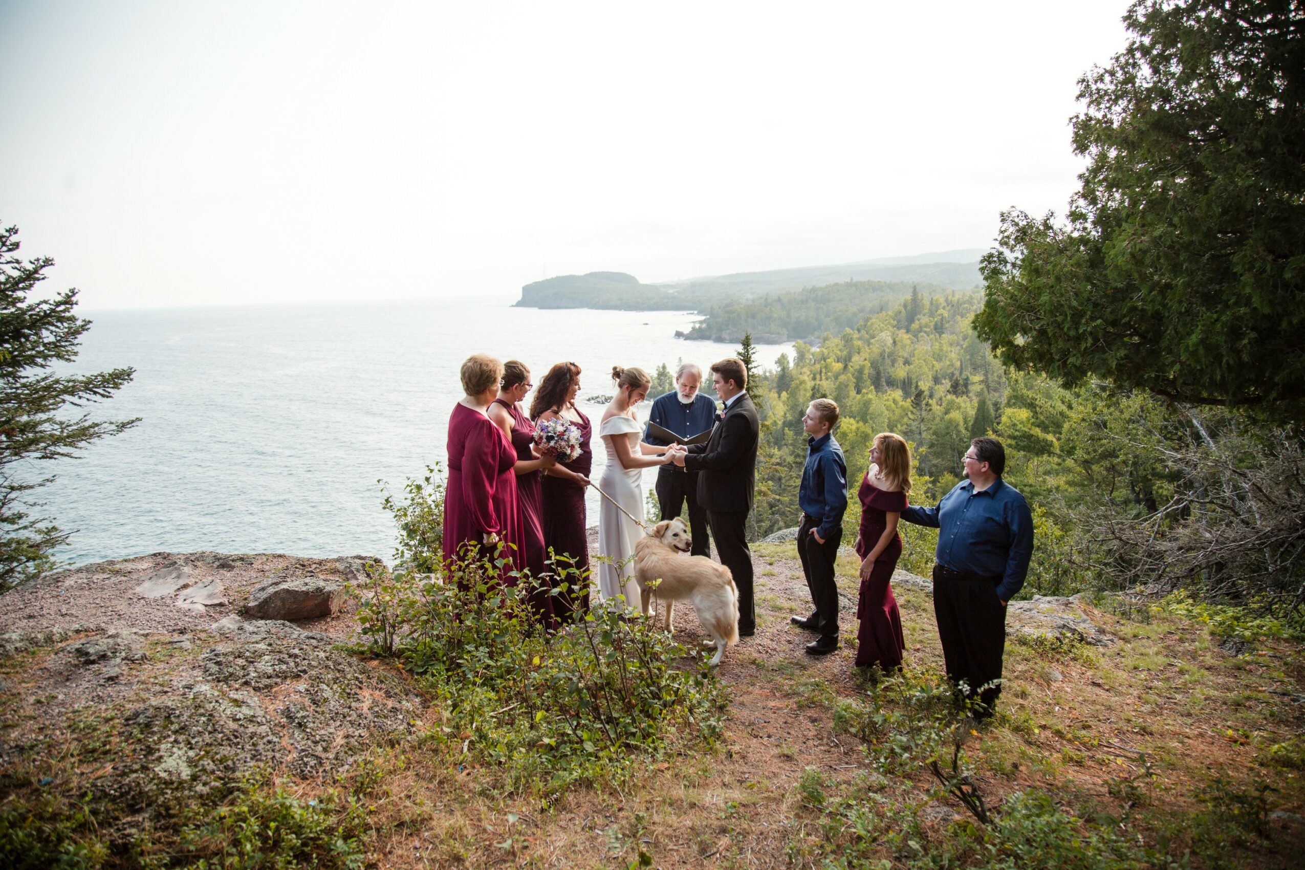 Group of people gathered outdoors near water, with trees and a distant shoreline in the background.