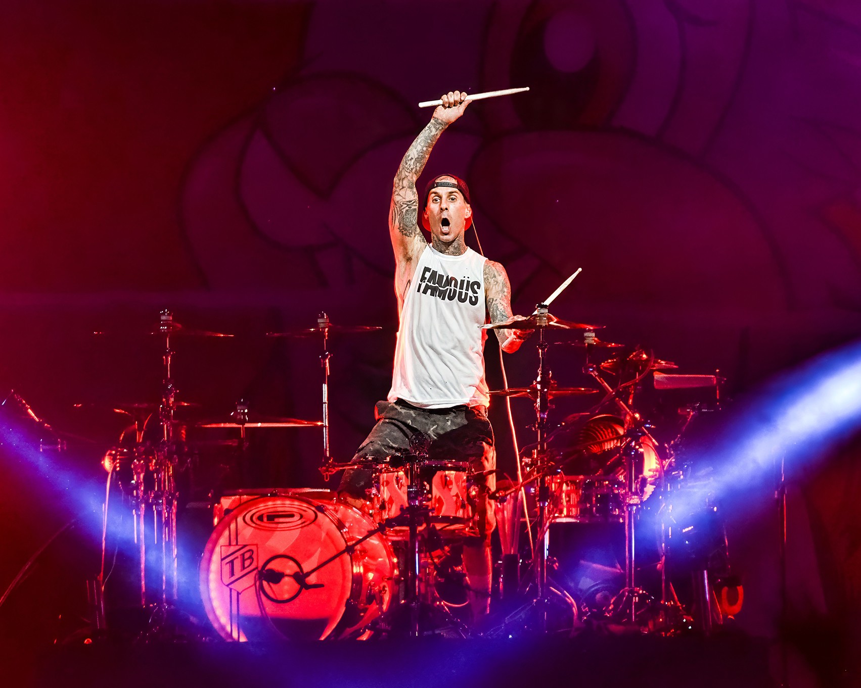 Performer with tattoos raising drumsticks on stage with drums and colorful lighting, wearing a white shirt with text.