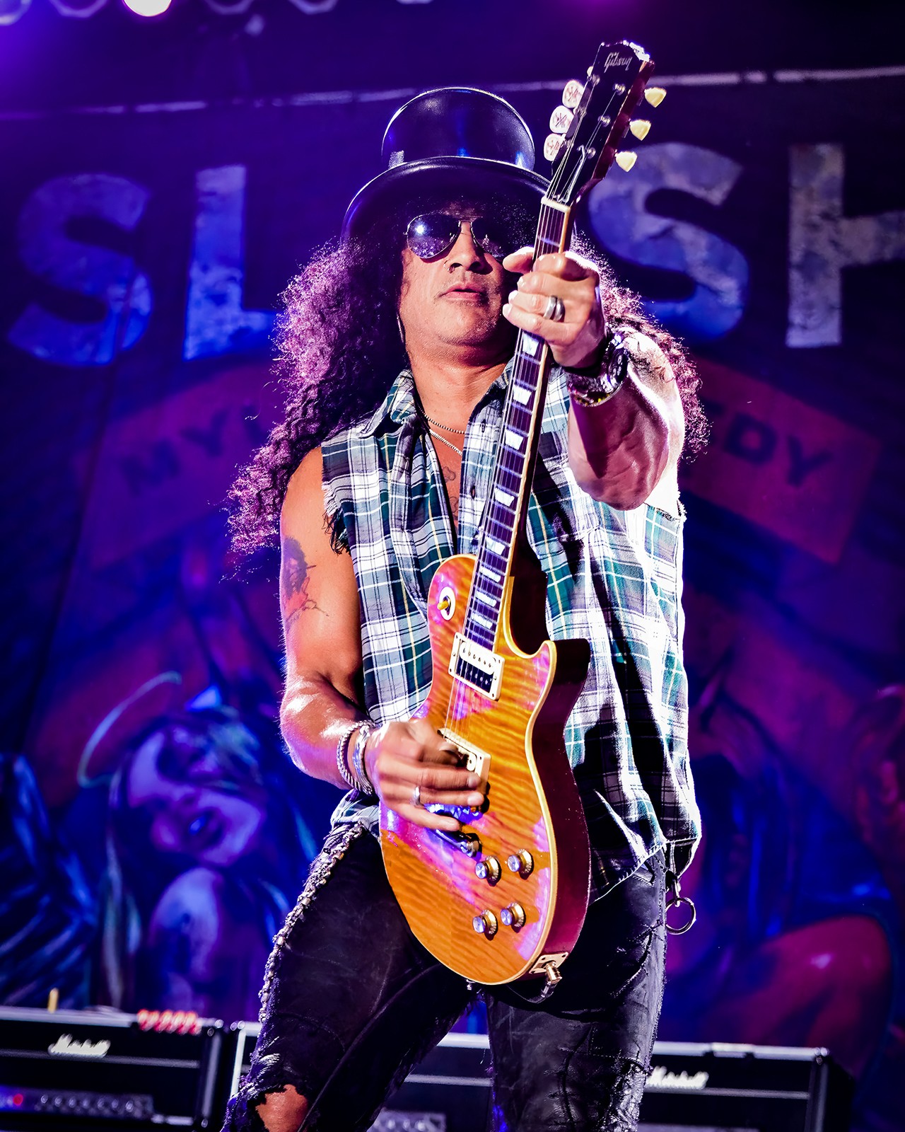 Person wearing sunglasses, a hat, and a sleeveless plaid shirt, holding an electric guitar on stage.