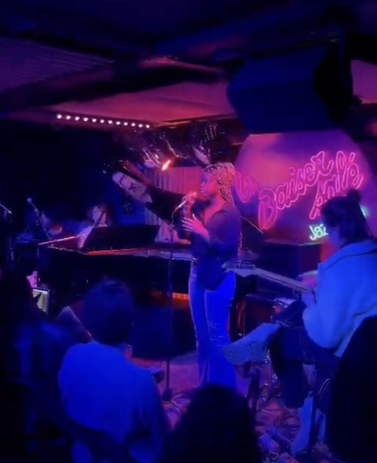 Person performing on stage with pink neon sign reading 'Fairy' in a dimly lit venue, audience watching attentively.