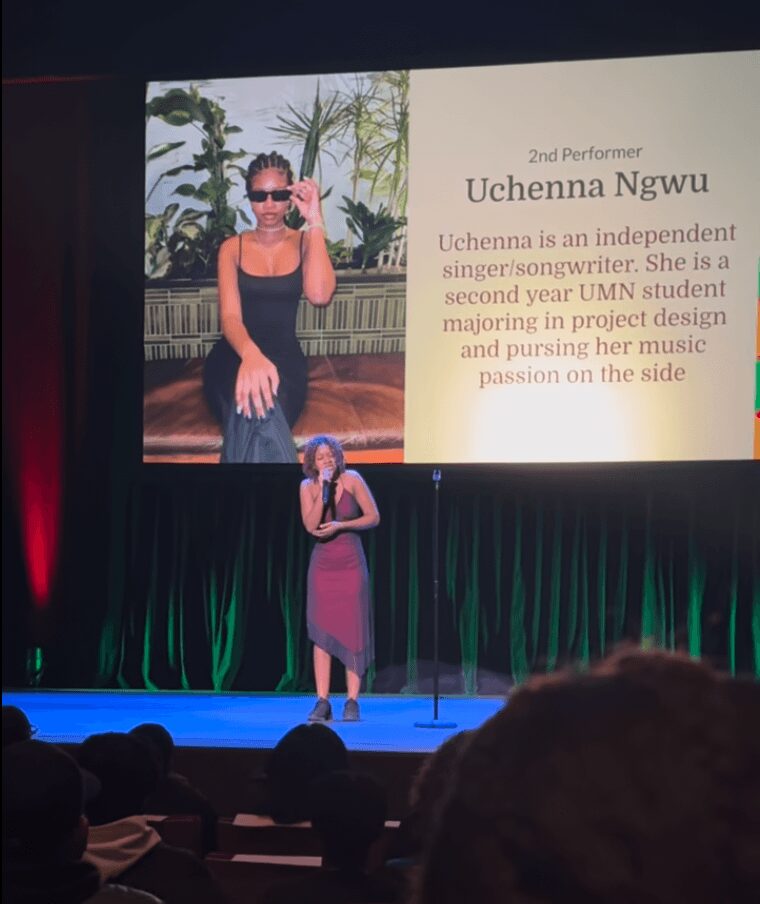 Woman in purple dress speaking on stage with large screen displaying her photo and biography behind her.