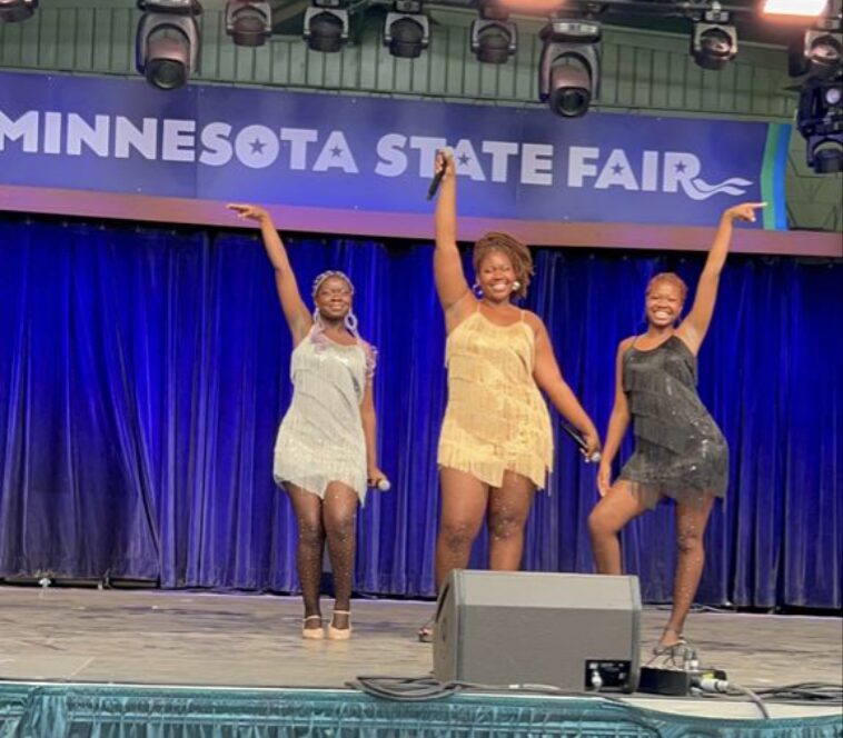 Three women in colorful dresses dancing on stage at the Minnesota State Fair, with a blue curtain background.