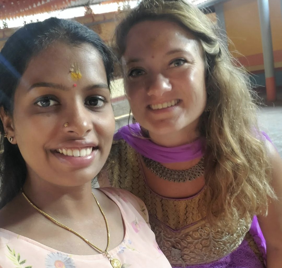 Two women smiling, one with traditional Indian attire and the other with a purple and gold outfit, in an indoor setting.