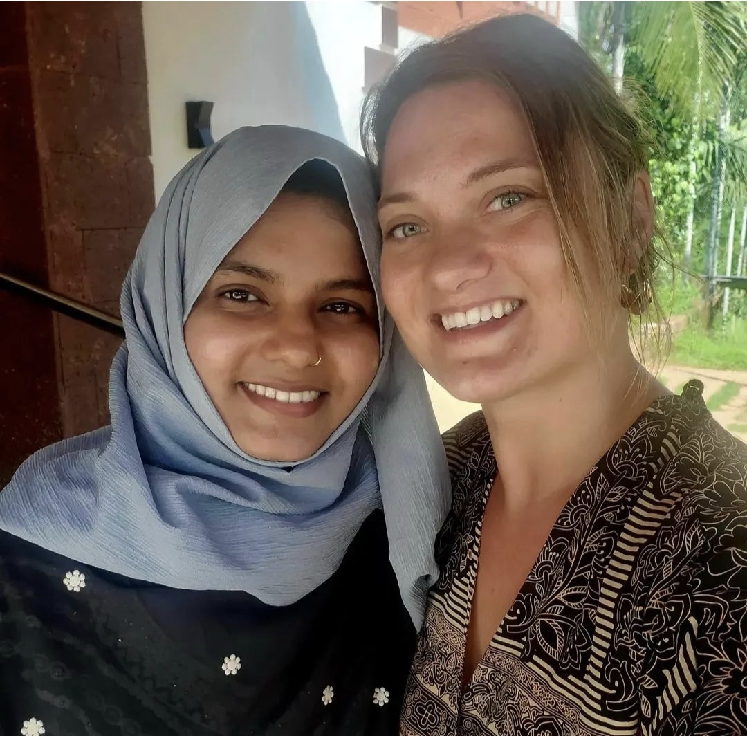 Two women smiling outdoors, one wearing a hijab and the other with light brown hair, in front of greenery.