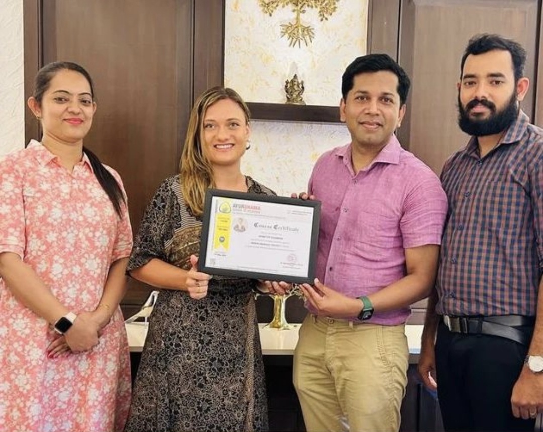 Four people standing indoors, one holding a framed certificate, smiling, with a decorative wall behind them.
