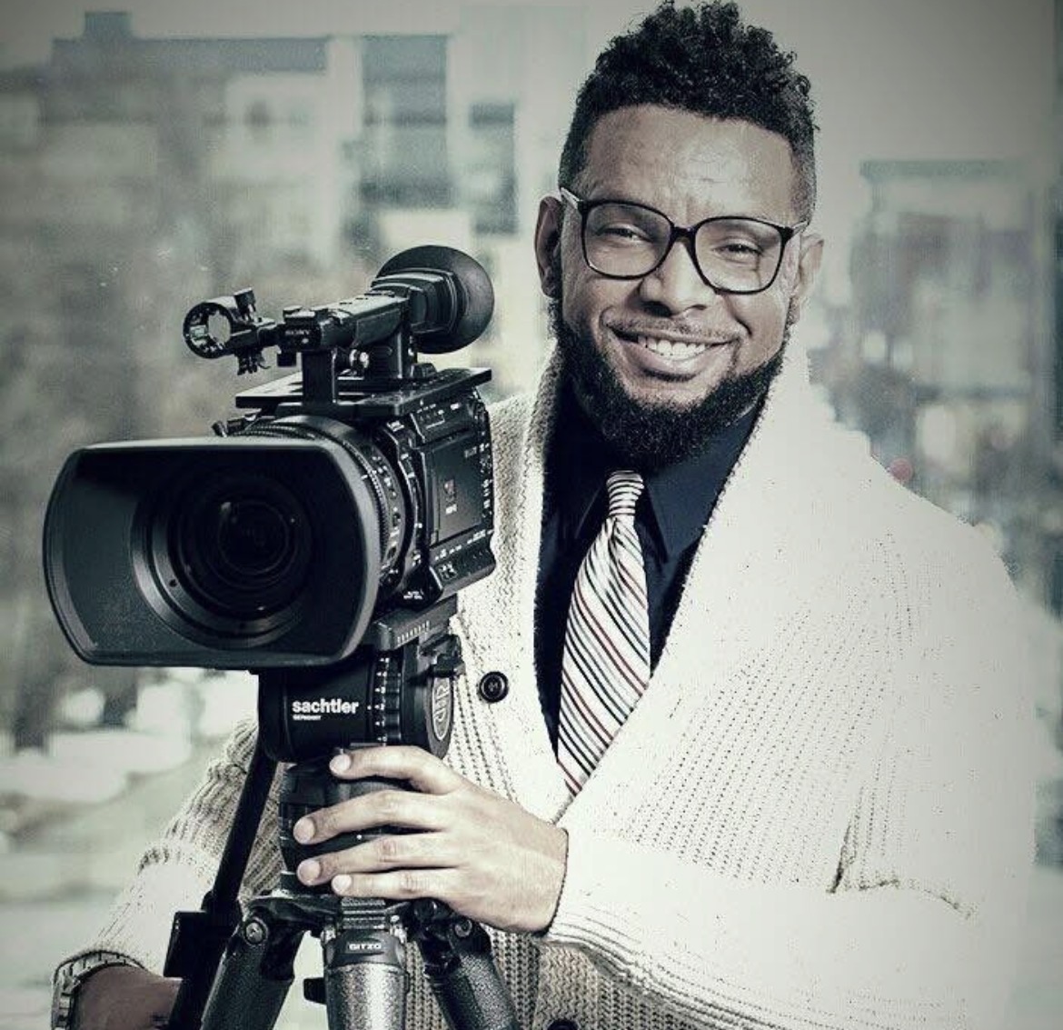 Man with glasses and a beard operating a professional video camera, smiling, in an indoor setting with windows in the background.