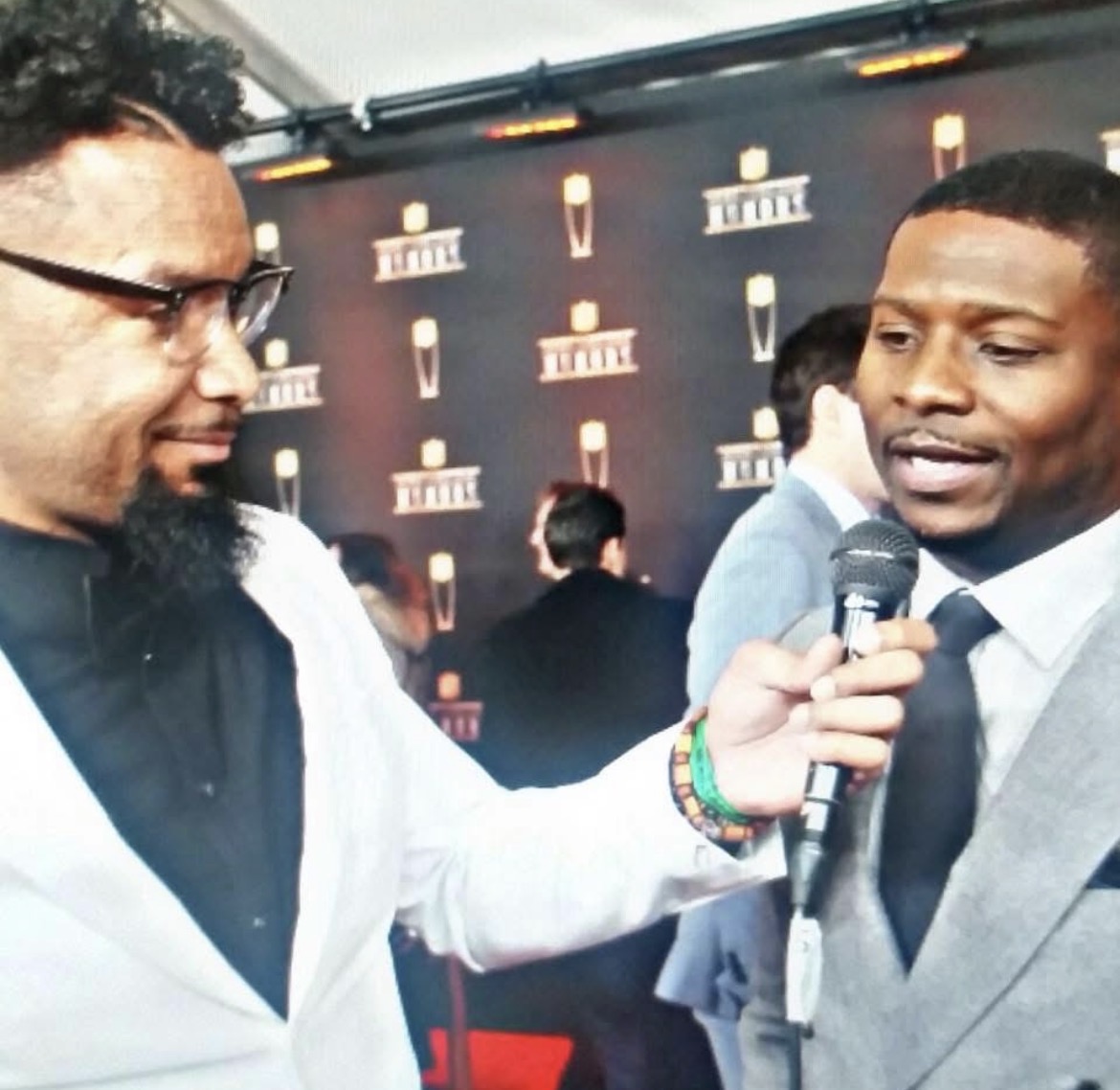 Two men engaged in an interview, one holding a microphone, at an event with a branded backdrop.