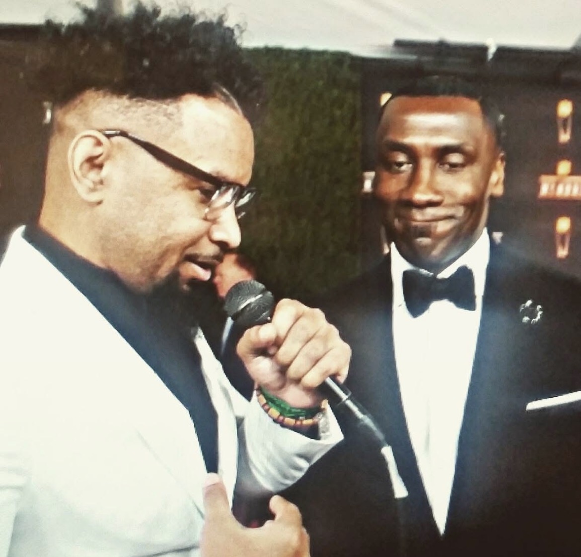 Two men in tuxedos, one speaking into a microphone, the other smiling, at a formal event.