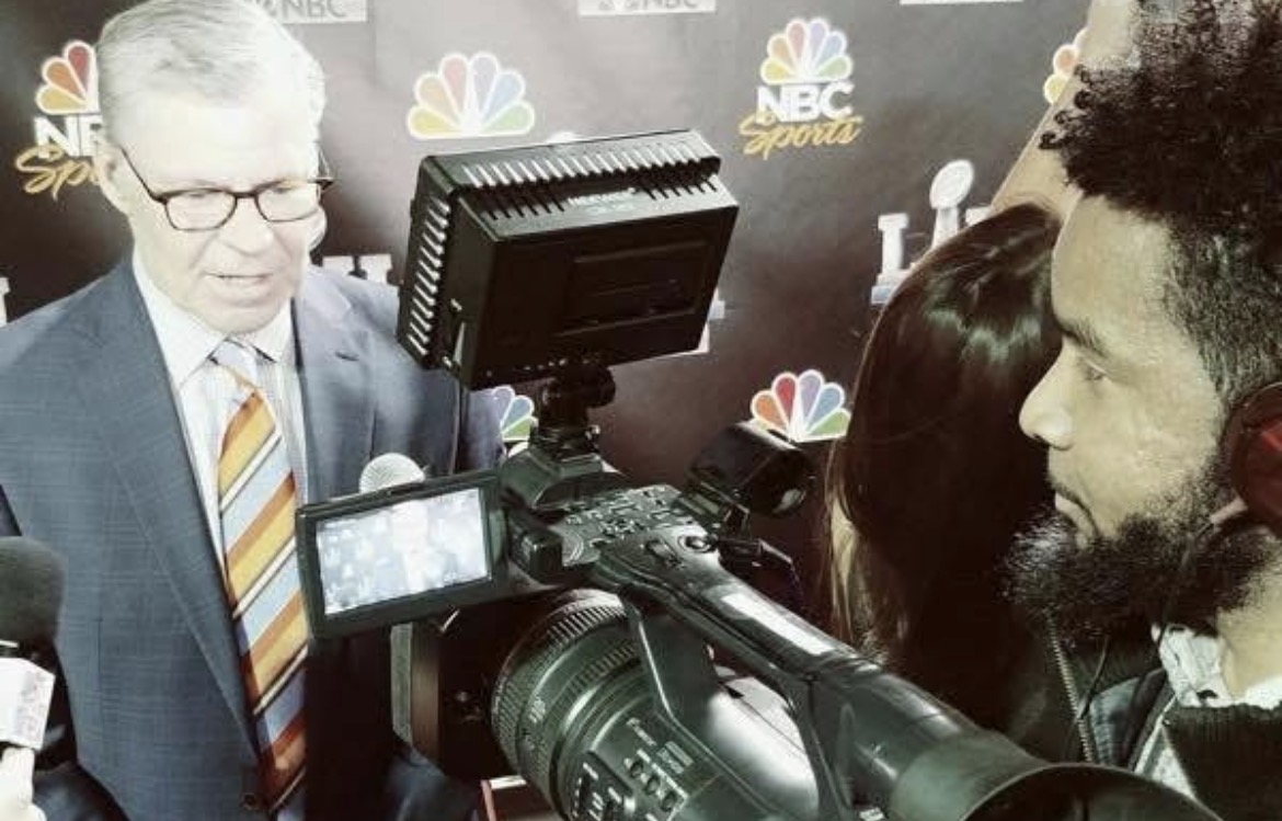 Man with glasses and striped tie being interviewed, camera and microphone in foreground, NBC Sports logo in background.