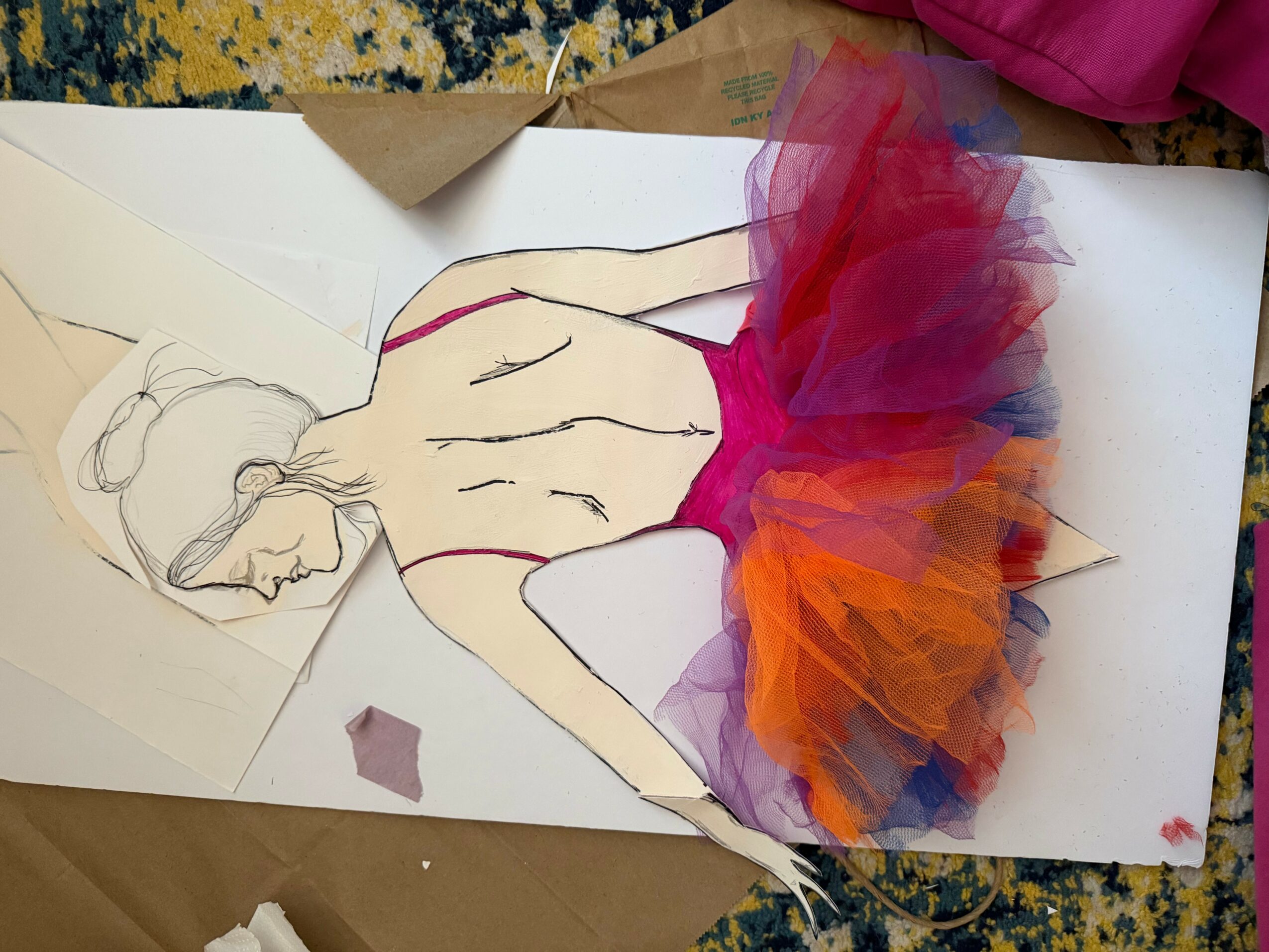 A paper cutout of a person with a colorful tutu, lying on a white surface with paper and fabric around.