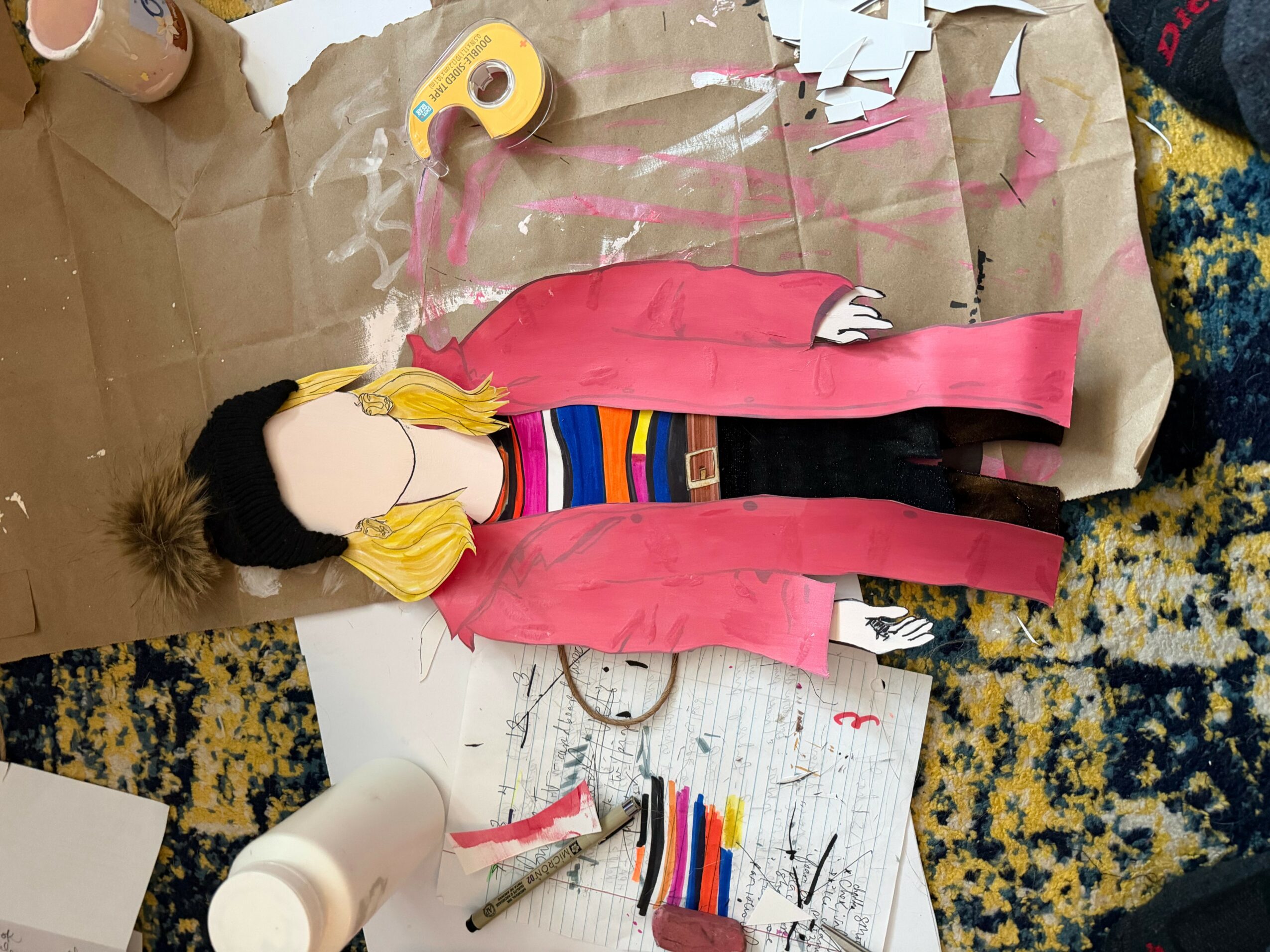 Person with long hair, pink jacket, and striped shirt working on paper crafts on a table.