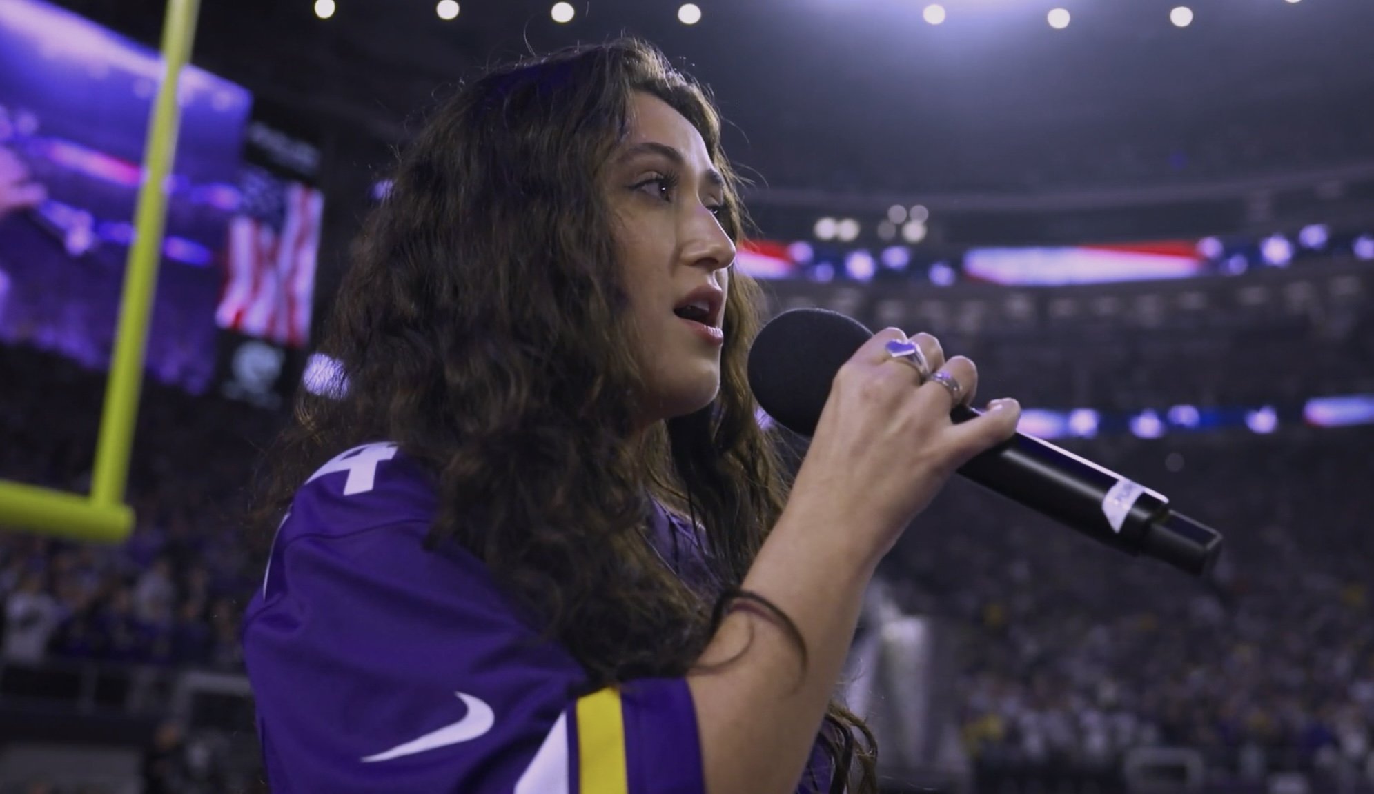 Young woman with long dark hair speaking into a microphone at a sports event, wearing a purple jersey.