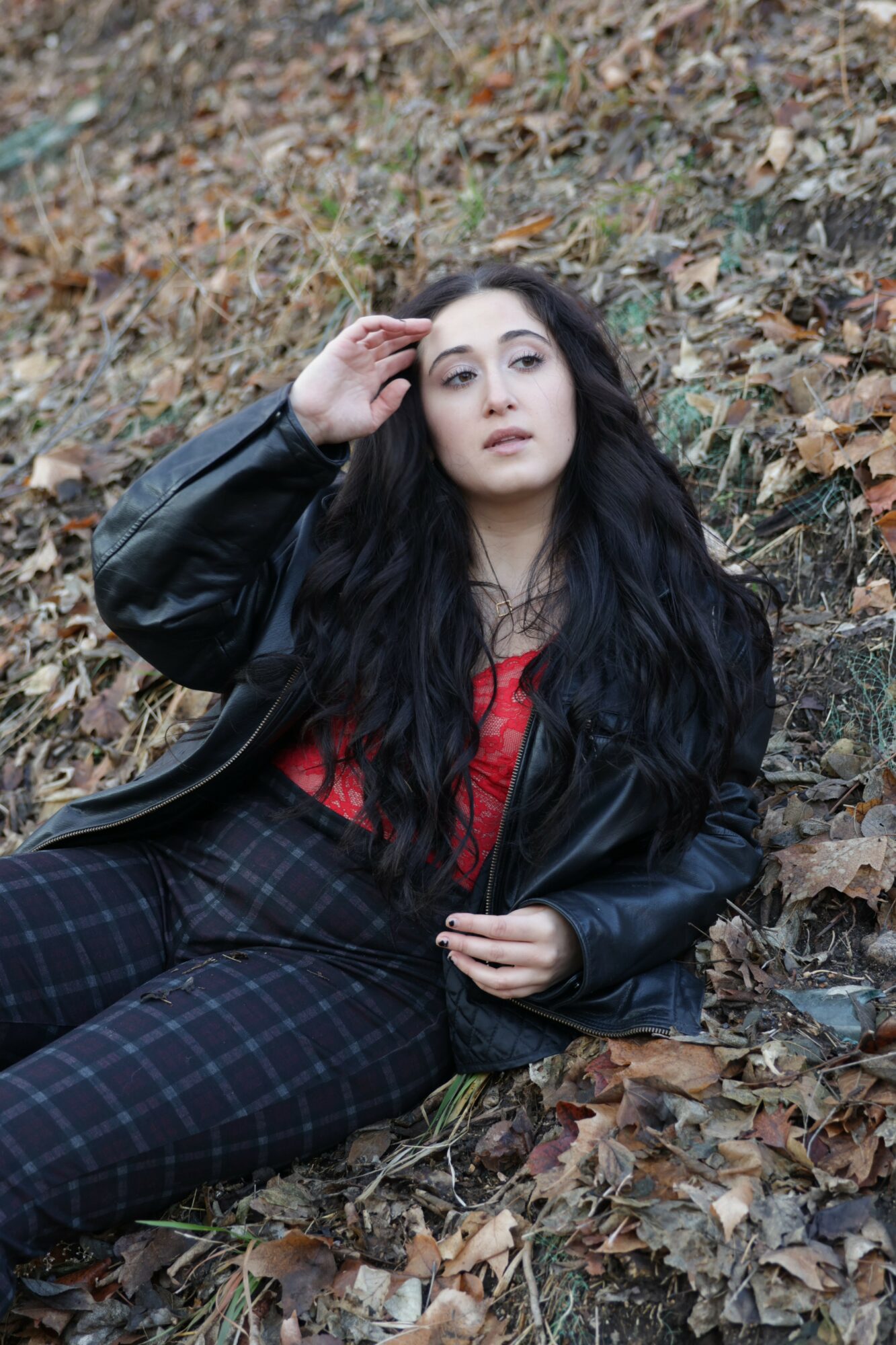 Young woman with long dark hair lying on fallen leaves outdoors, wearing a black leather jacket and red top.