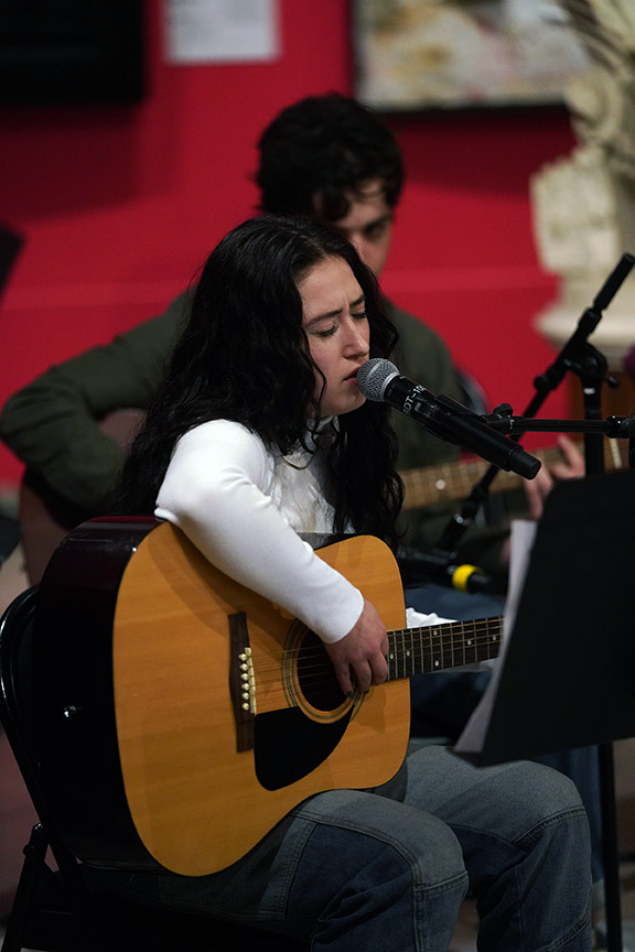 Woman with dark hair playing guitar and singing into a microphone, with a person in the background, in a red room.