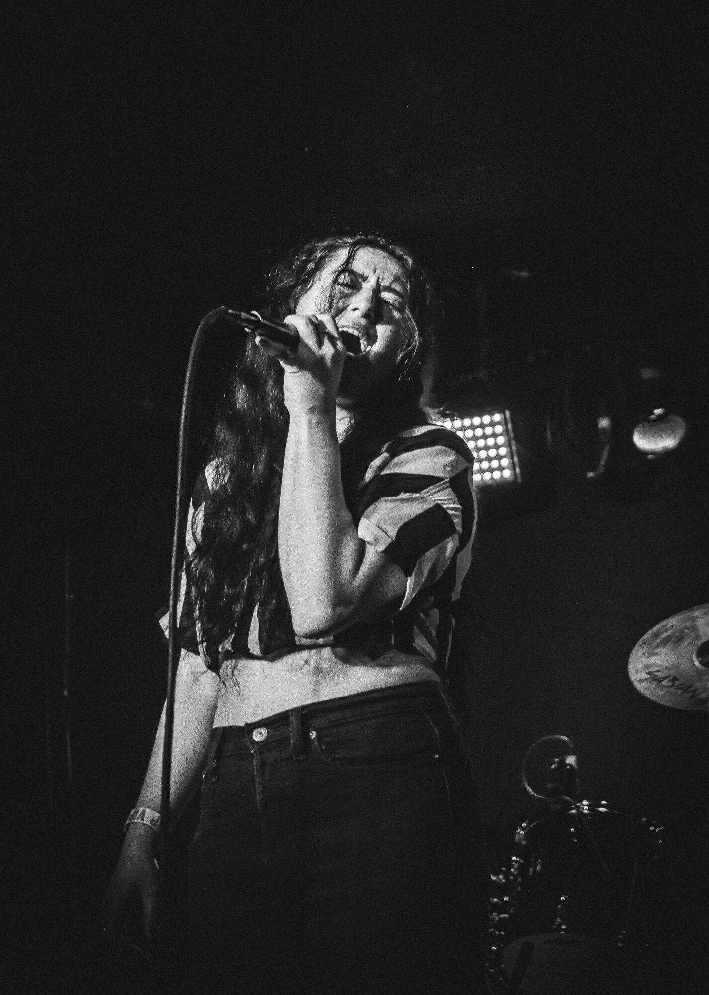 Person singing into a microphone on stage with dark background, wearing striped shirt and high-waisted pants.