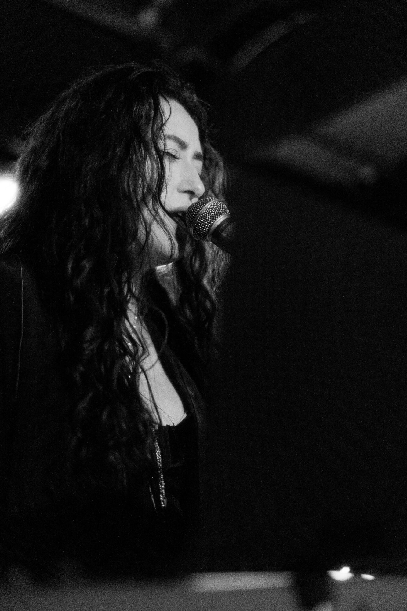Woman singing into a microphone with long curly hair, in a dark setting.