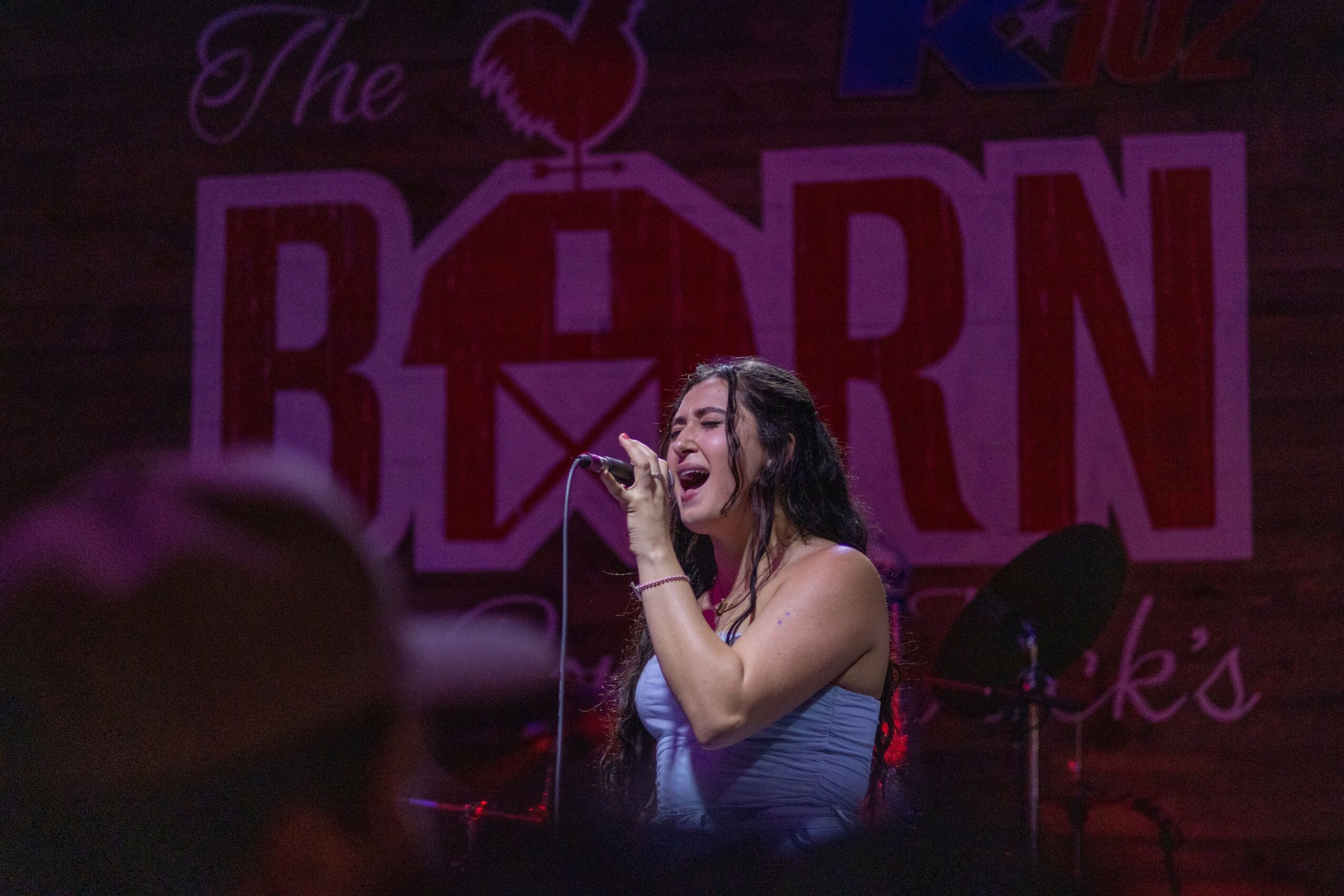 A woman singing into a microphone on stage with a pink and purple background, holding a hand near her face.