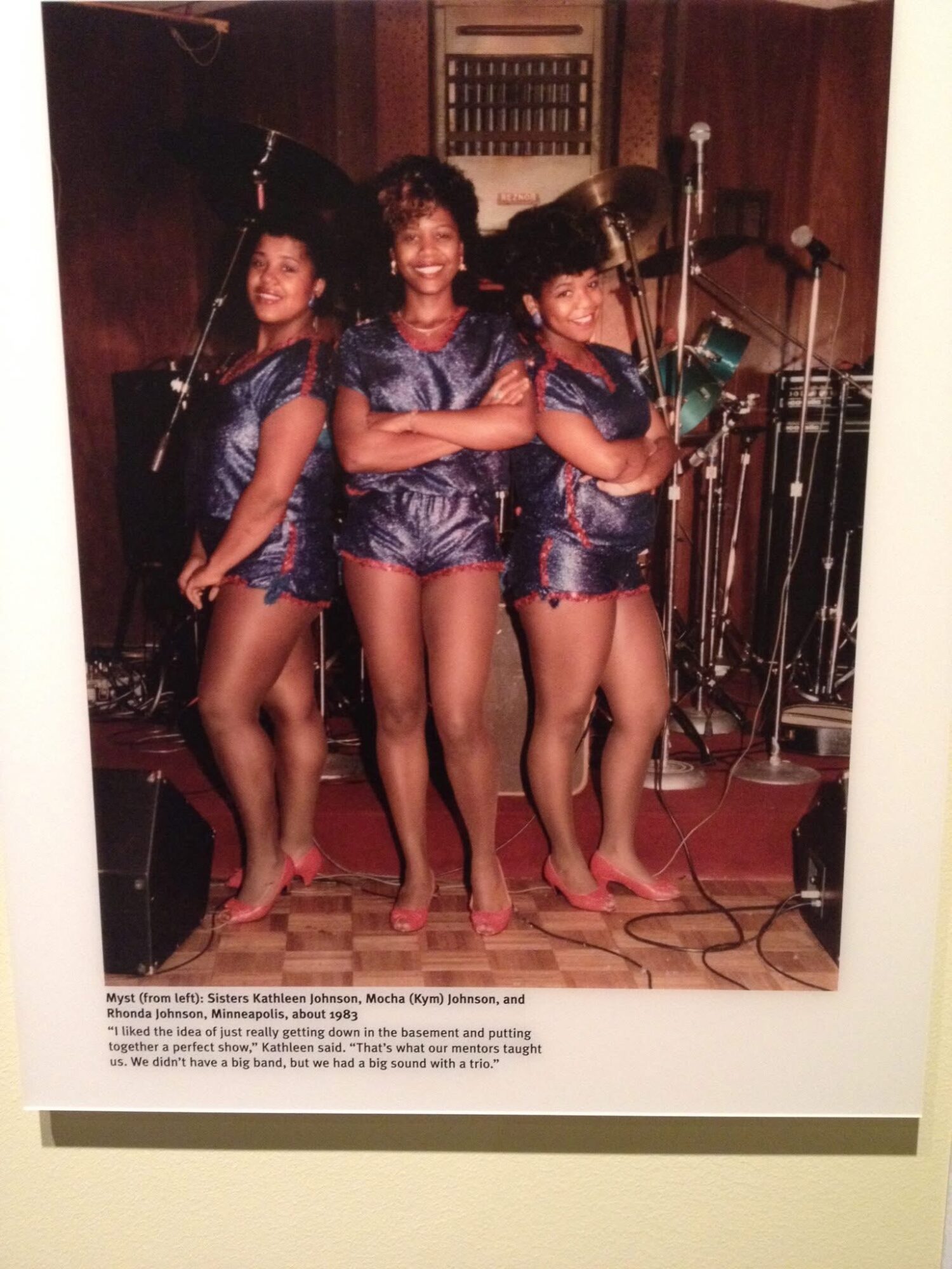 Three young girls in matching shiny blue costumes pose on stage with microphones and musical equipment behind them.