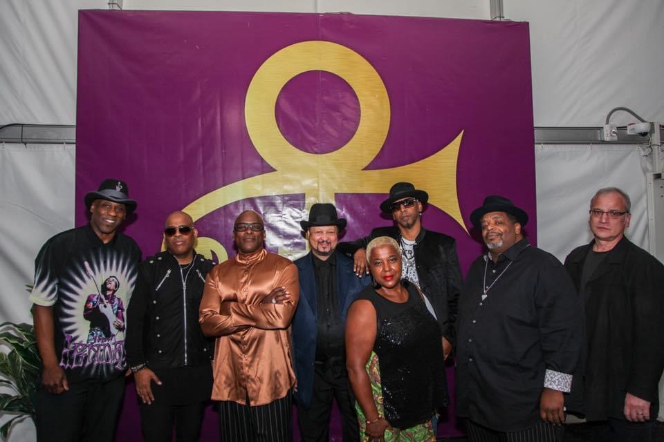 Group of eight people standing in front of a purple backdrop with a yellow anchor symbol, smiling and posing for the photo.