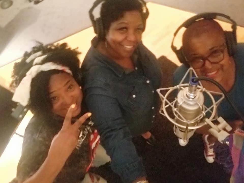 Three people in a recording studio, smiling, with a microphone and headphones, posing for the photo.