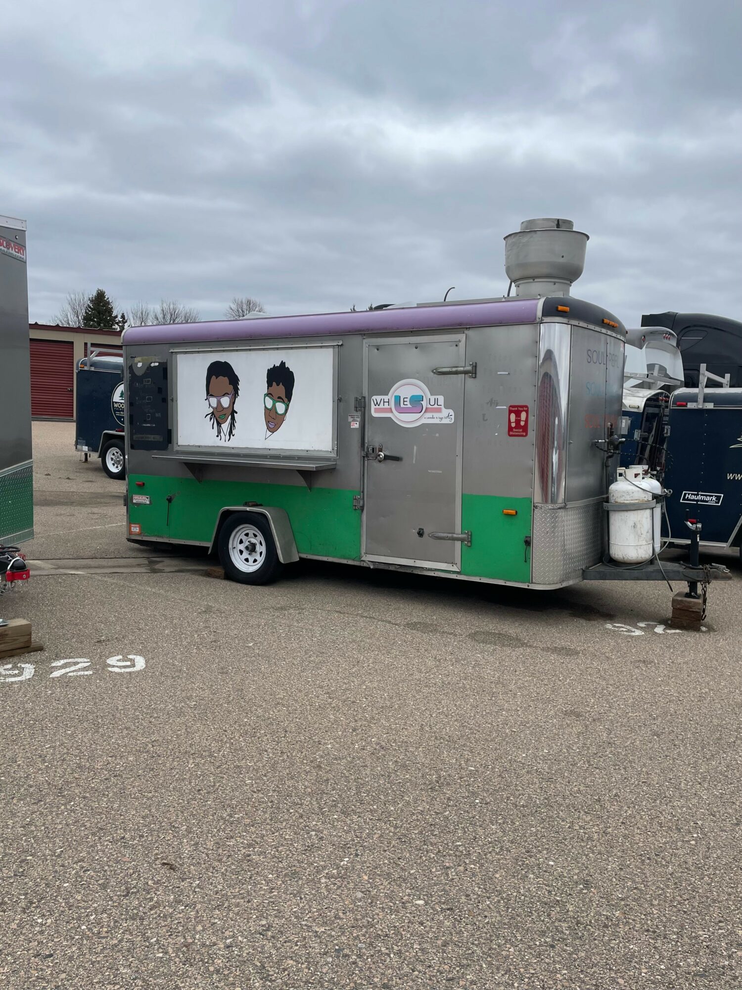 A silver and green food trailer with images of two people on the side, parked in a lot under cloudy sky.